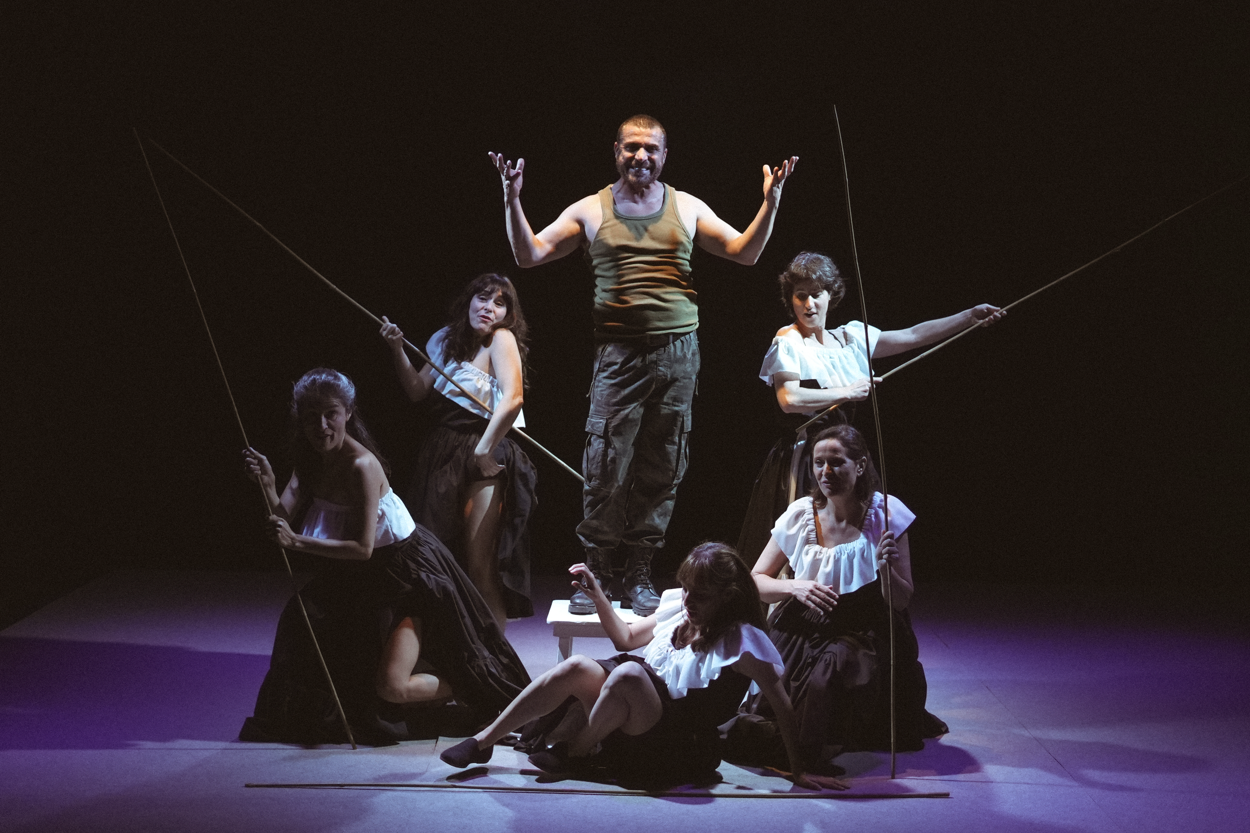 Un grupo de seis actores en el escenario durante una obra de teatro con un fondo oscuro, uno de ellos de pie en el centro con una expresión de alegría y brazos levantados, rodeados de cinco actores femeninas en diferentes posiciones, usando ropa de estilo rústico y sosteniendo varas, en una puesta en escena teatral.