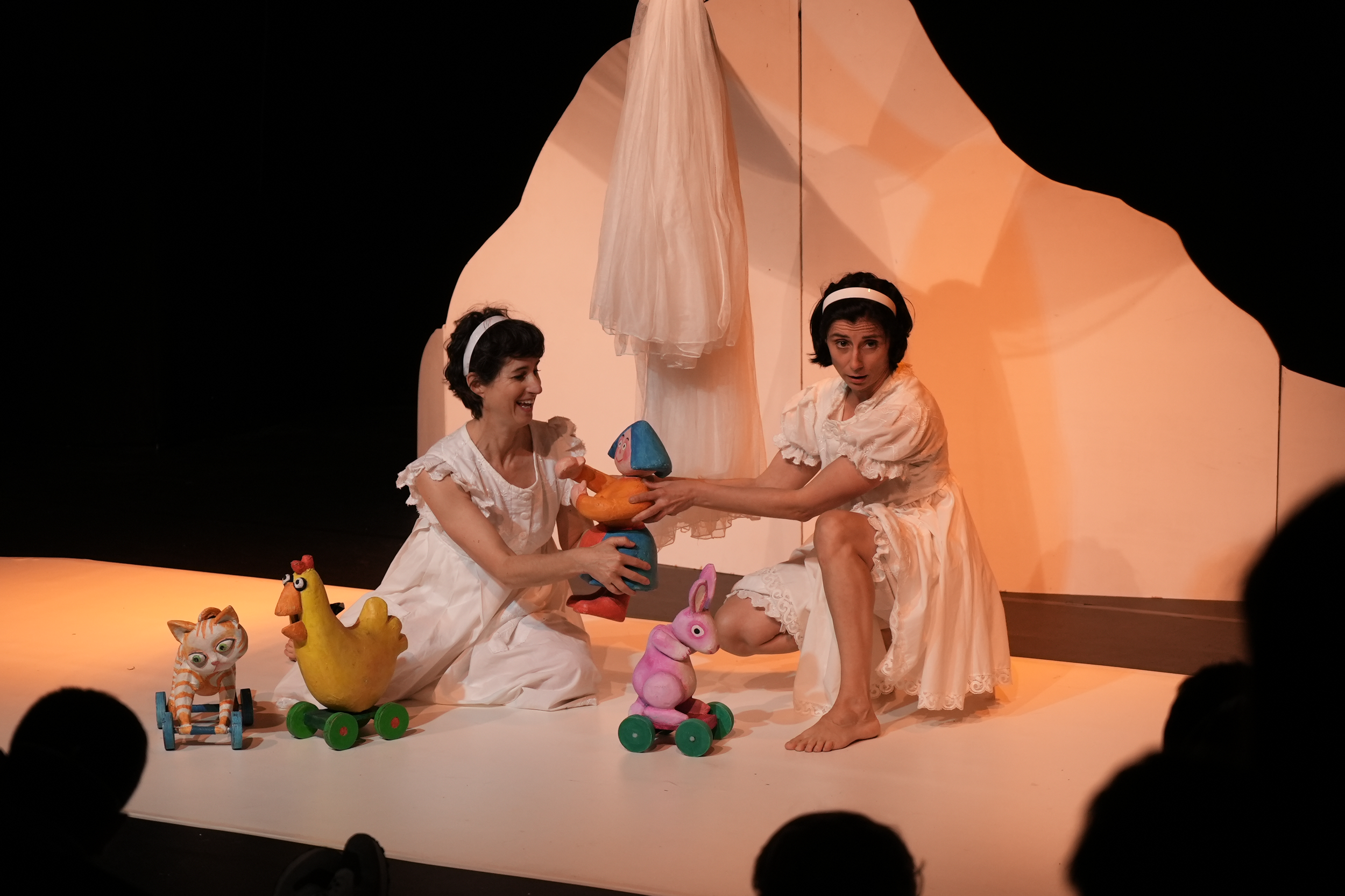 Dos mujeres con vestimenta blanca en un escenario negro, sosteniendo títeres de animales y personajes de juguetes en un teatro infantil.