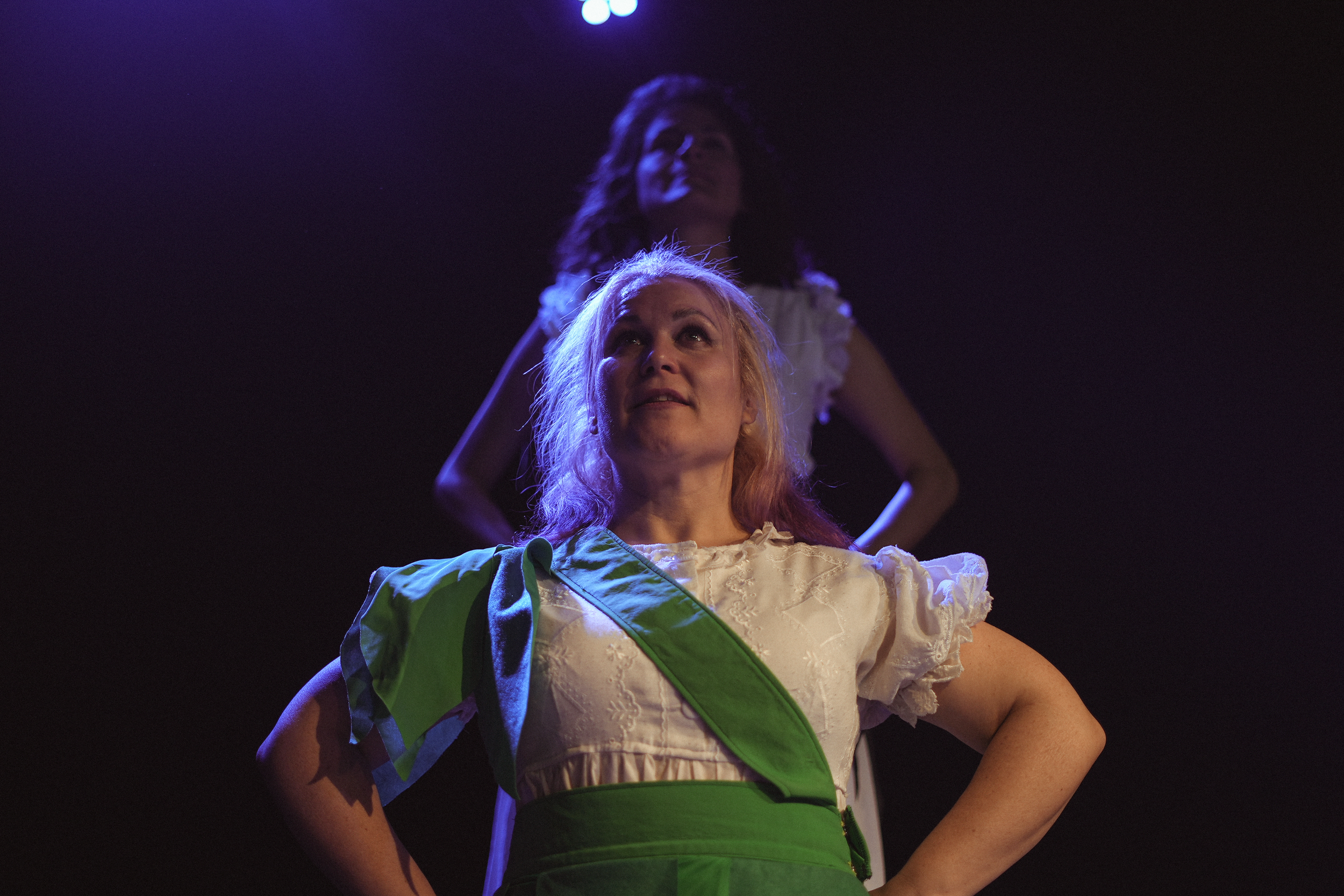 Dos mujeres en un escenario con iluminación teatral, una en primer plano con expresión orgullosa y la otra al fondo con las manos en la cintura, ambas vestidas con ropa vintage en un escenario oscuro.