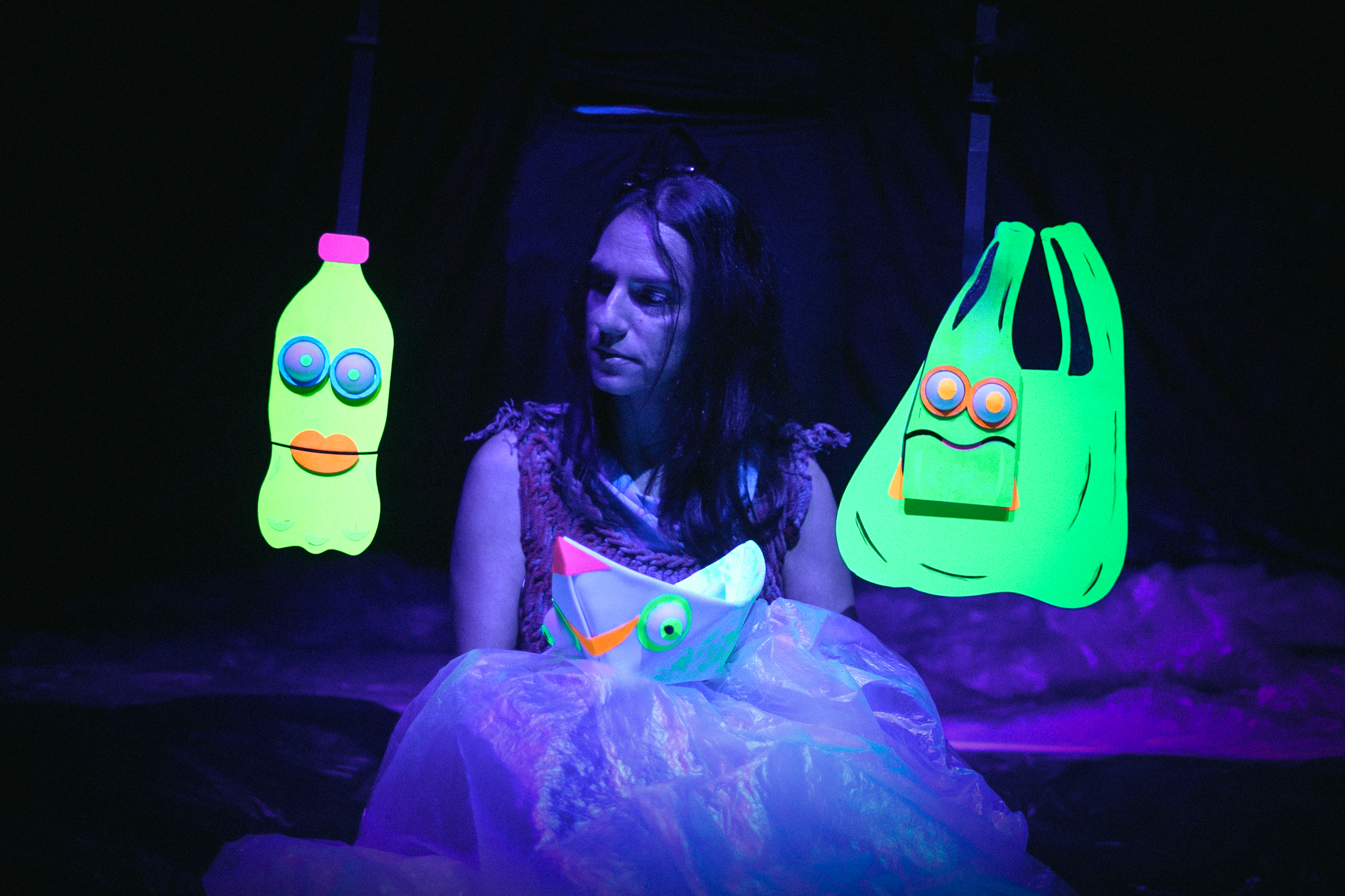 Una mujer sentada en un escenario oscuro con decoraciones brillantes y fluorescentes, incluyendo figuras de bolsas y una botella, con efectos de luz negra.