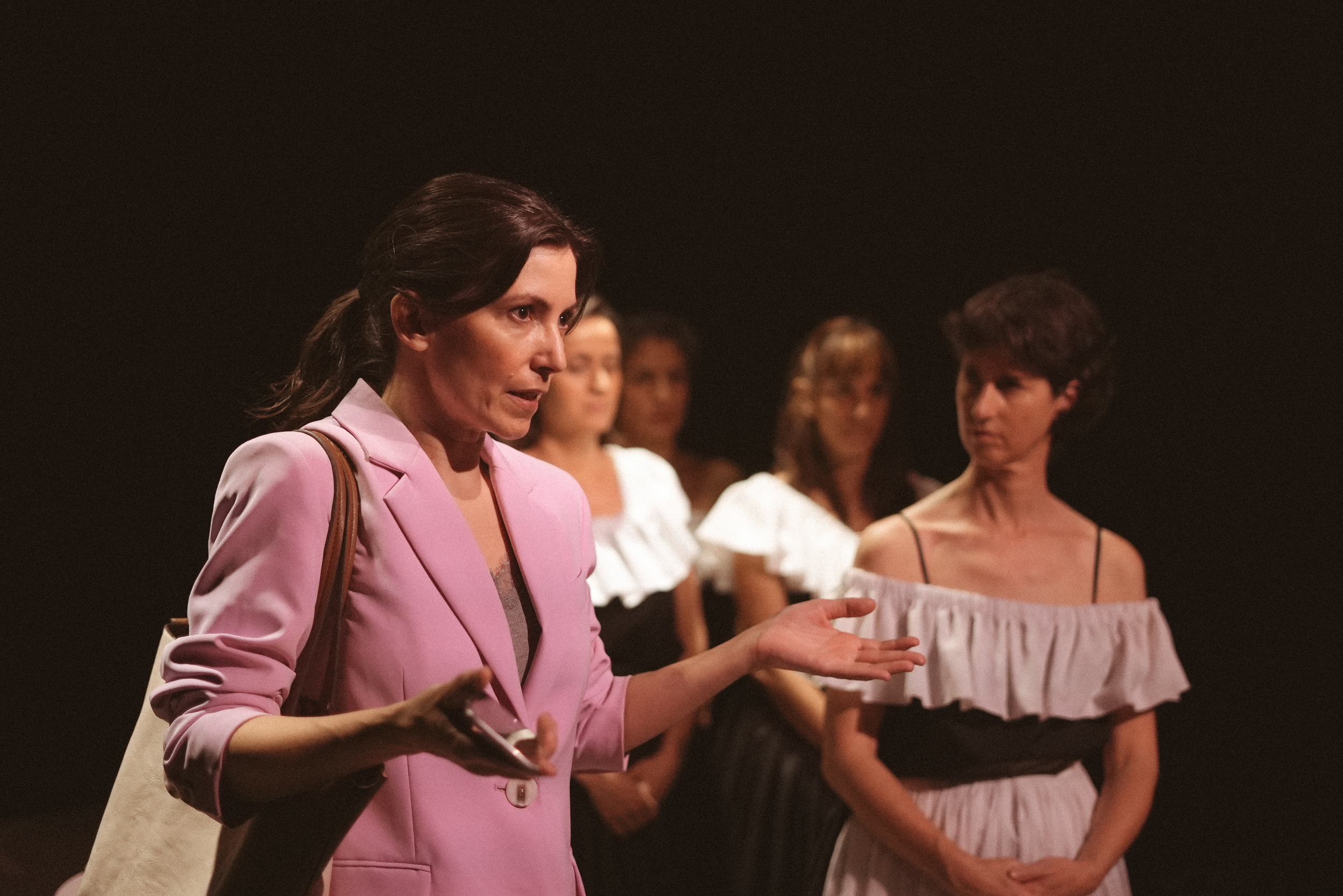Mujer con blazer rosa hablando, acompañada por otras mujeres en el fondo, en un escenario oscuro.