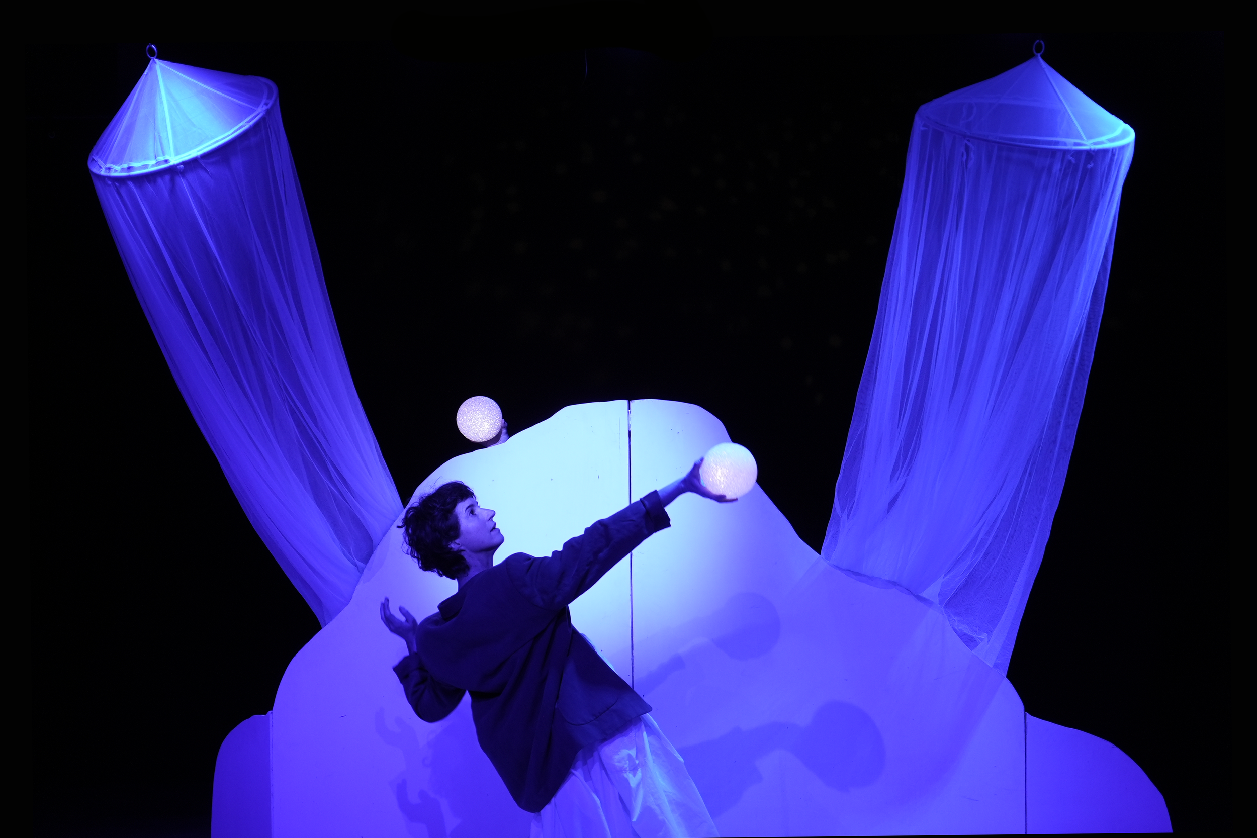 Persona que realiza un acto teatral o artístico en un escenario con iluminación azul, con un fondo oscuro y estructuras de tela y luz.