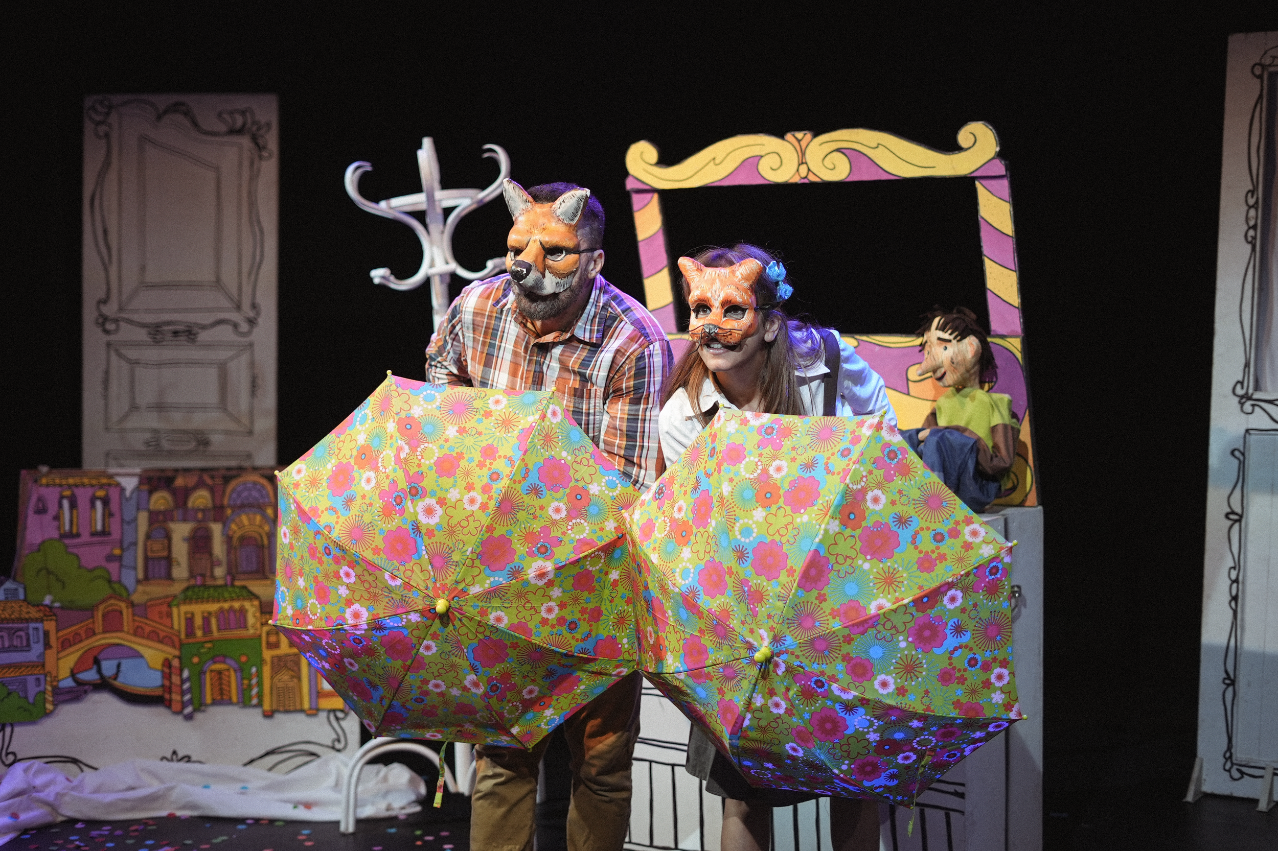 Dos actores con máscaras de animales en un escenario teatral, sosteniendo paraguas con patrones coloridos y fondo pintado con elementos de una ciudad y personajes de teatro.