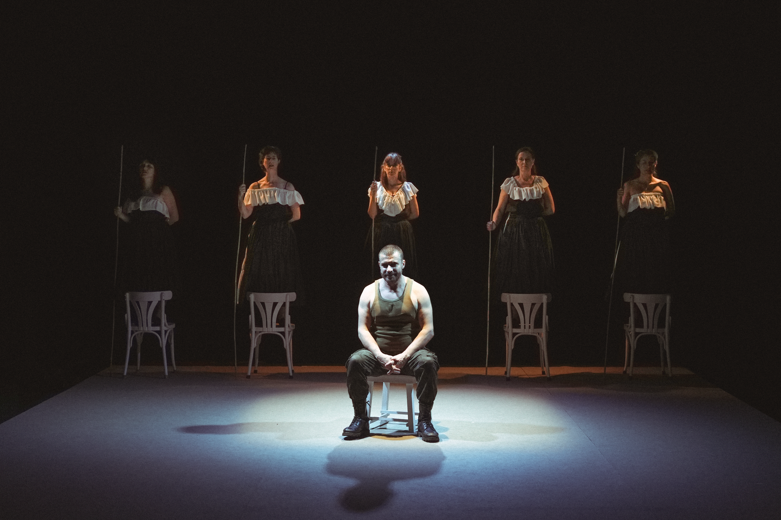 Un grupo de cinco personas en un escenario oscuro, una de ellas sentada en una silla en el centro y las demás de pie con bastones, en lo que parece ser una escena teatral.