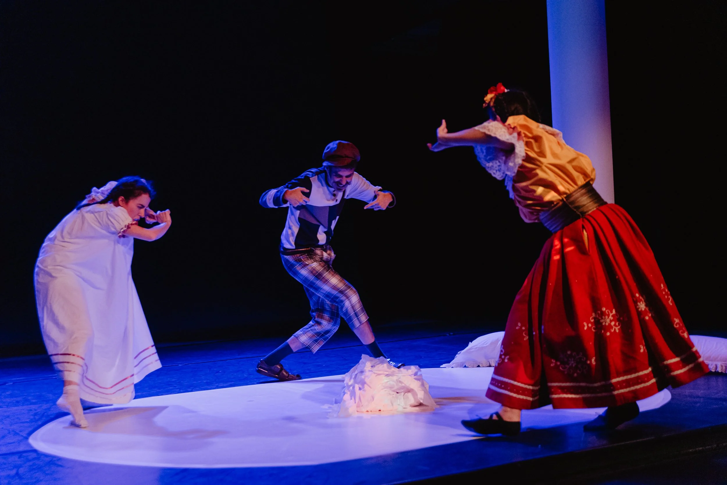 Tres actores en escena durante una obra teatral, disfrazados con ropa tradicional, con uno en el centro en una posición dinámica y los otros dos a los lados en diferentes gestos.