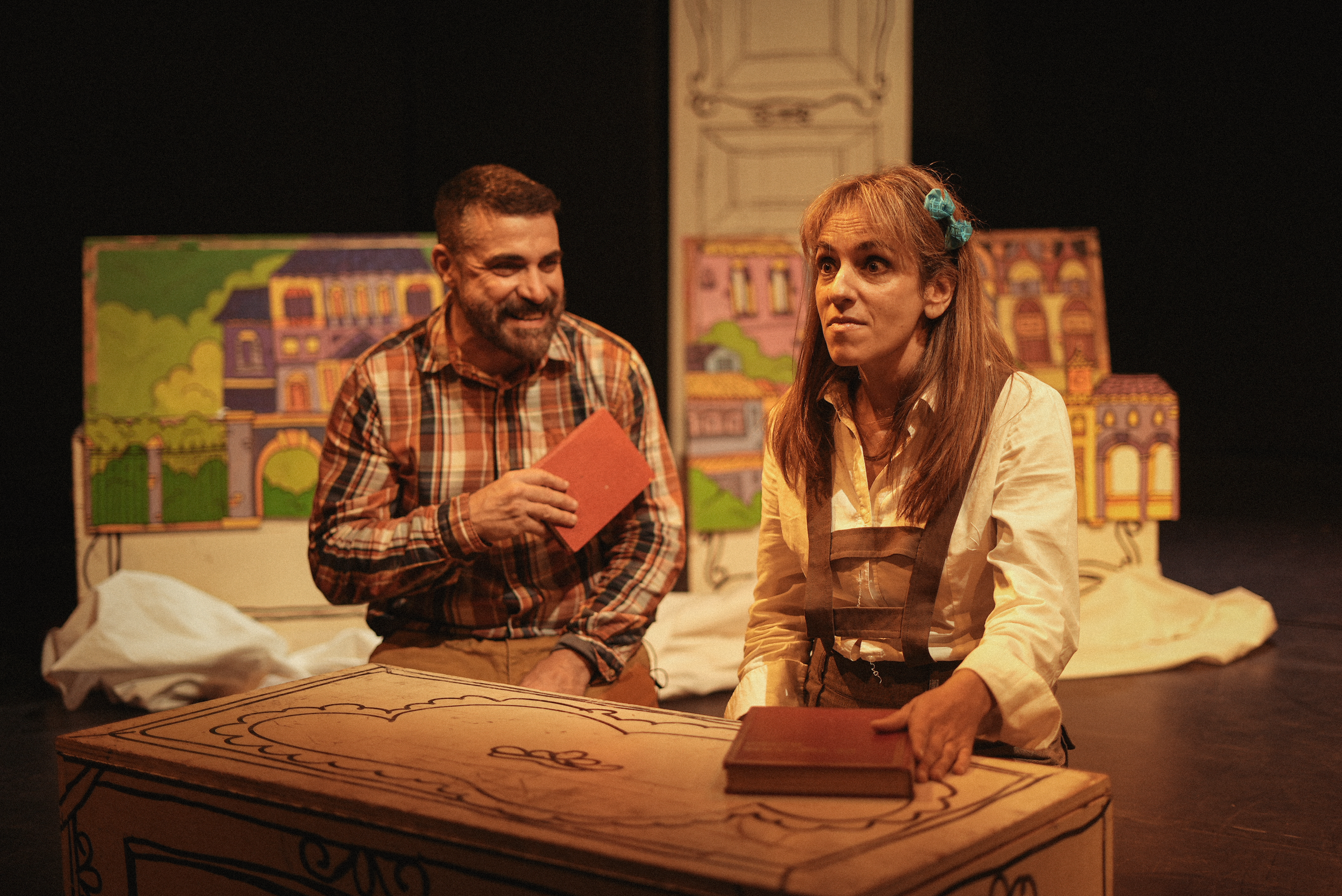 Dos actores en un escenario teatral con decorados de casas y árboles pintados, uno de ellos con una expresión sonriente y el otro con expresión pensativa, ambos rodeados de muebles y accesorios como libros y una mesa pintada.