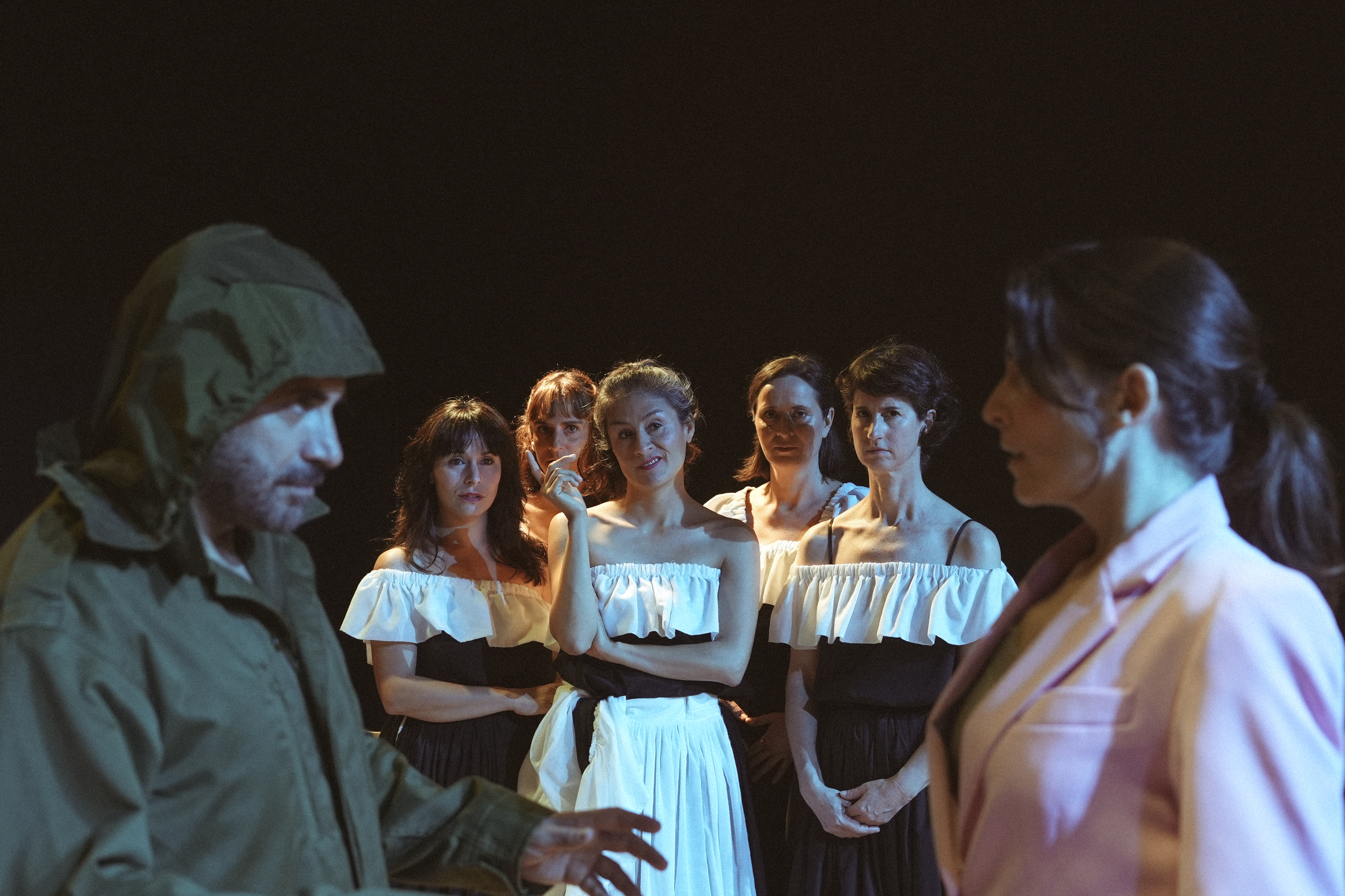 Una escena teatral con siete personajes en un escenario oscuro, vestidos con ropa que recuerda trajes tradicionales de país latinoamericano, en una conversación seria