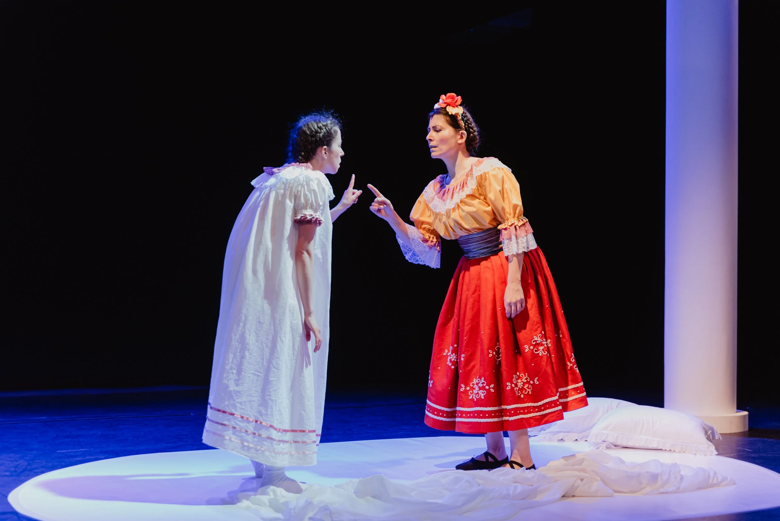 Dos mujeres en un escenario teatral, una vestida de blanco y la otra con un vestido rojo y blusa amarilla, discutiendo con gestos intensos, en un escenario con fondo oscuro y una columna blanca a la derecha.