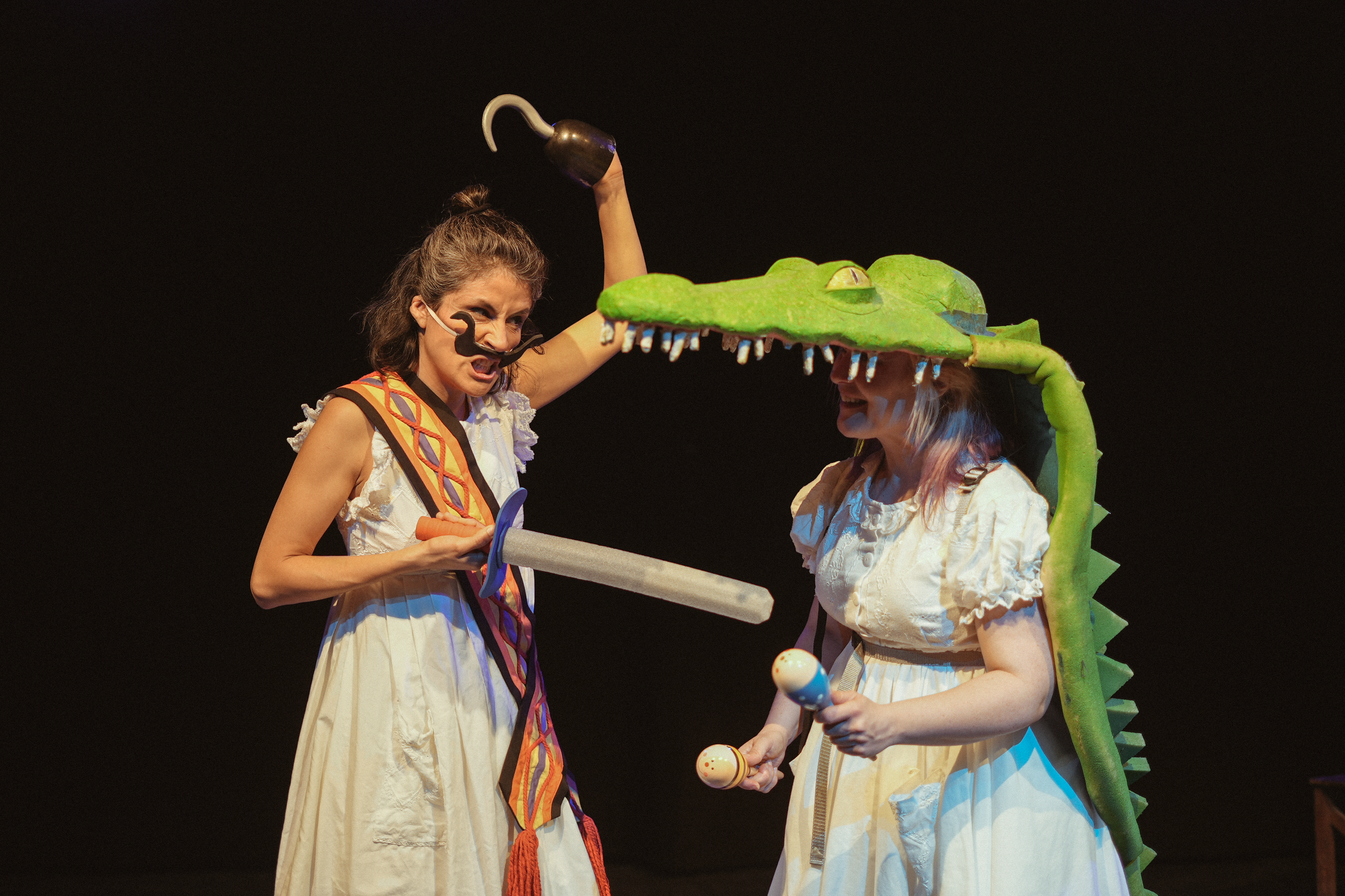 Dos personas disfrazadas de personajes de teatro, una con un sombrero de cocodrilo y otra con una espada de madera y maracas, en un escenario oscuro.