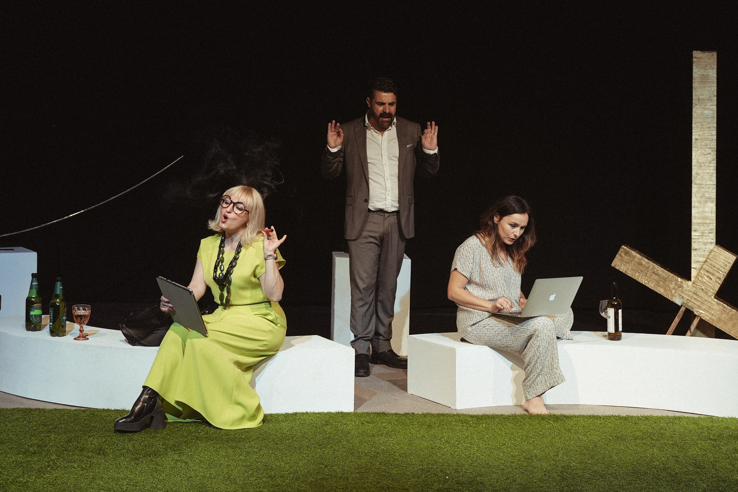 Tres personas en escena, una mujer con vestido amarillo y gafas, otra mujer con pijama y laptop, y un hombre en medio con traje, en un escenario minimalista con estructuras de madera y objetos sobre una plataforma blanca.
