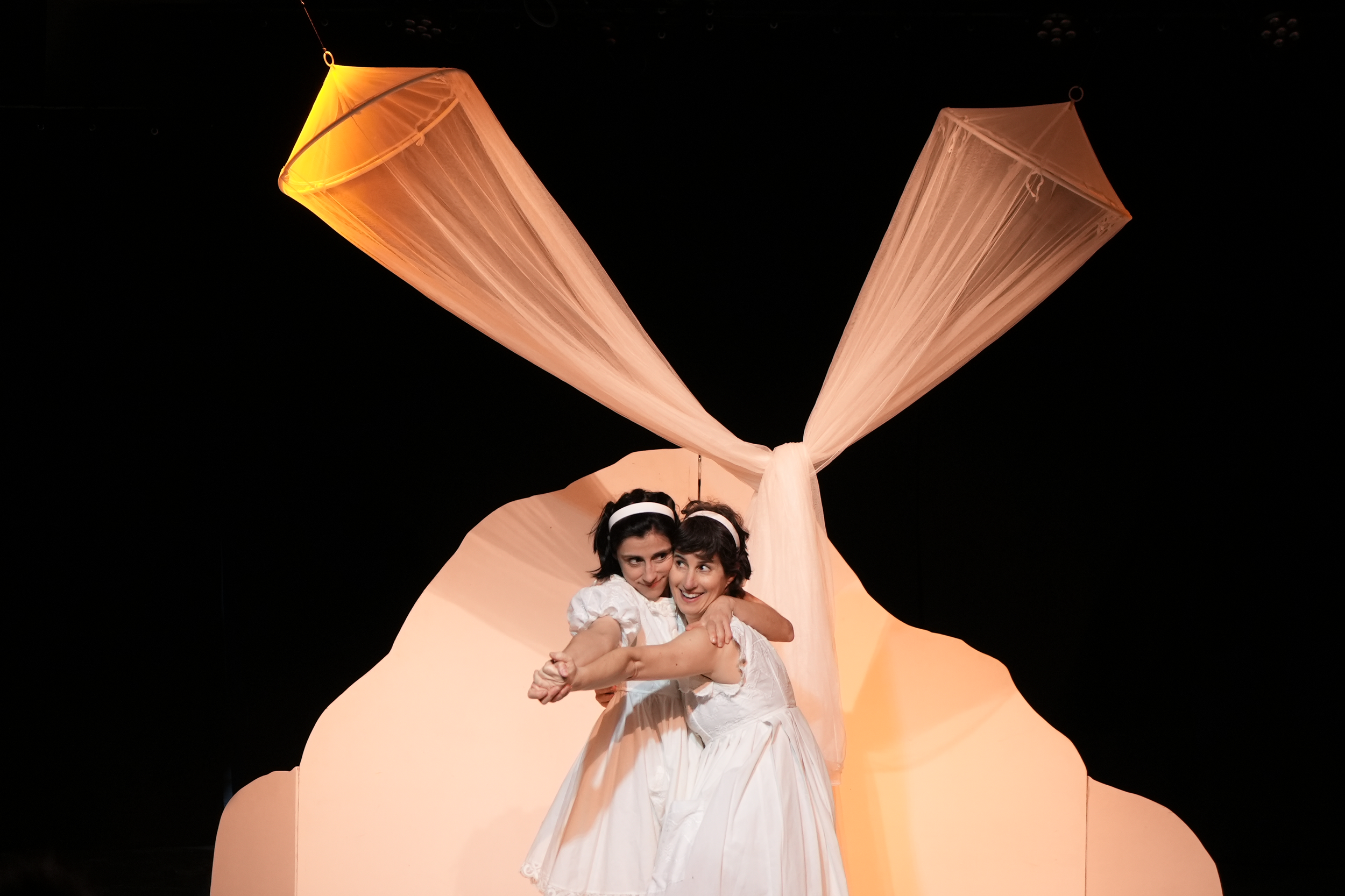 Dos mujeres en vestuarios blancos en escena teatral, con cortinas blancas en forma de mariposa de fondo