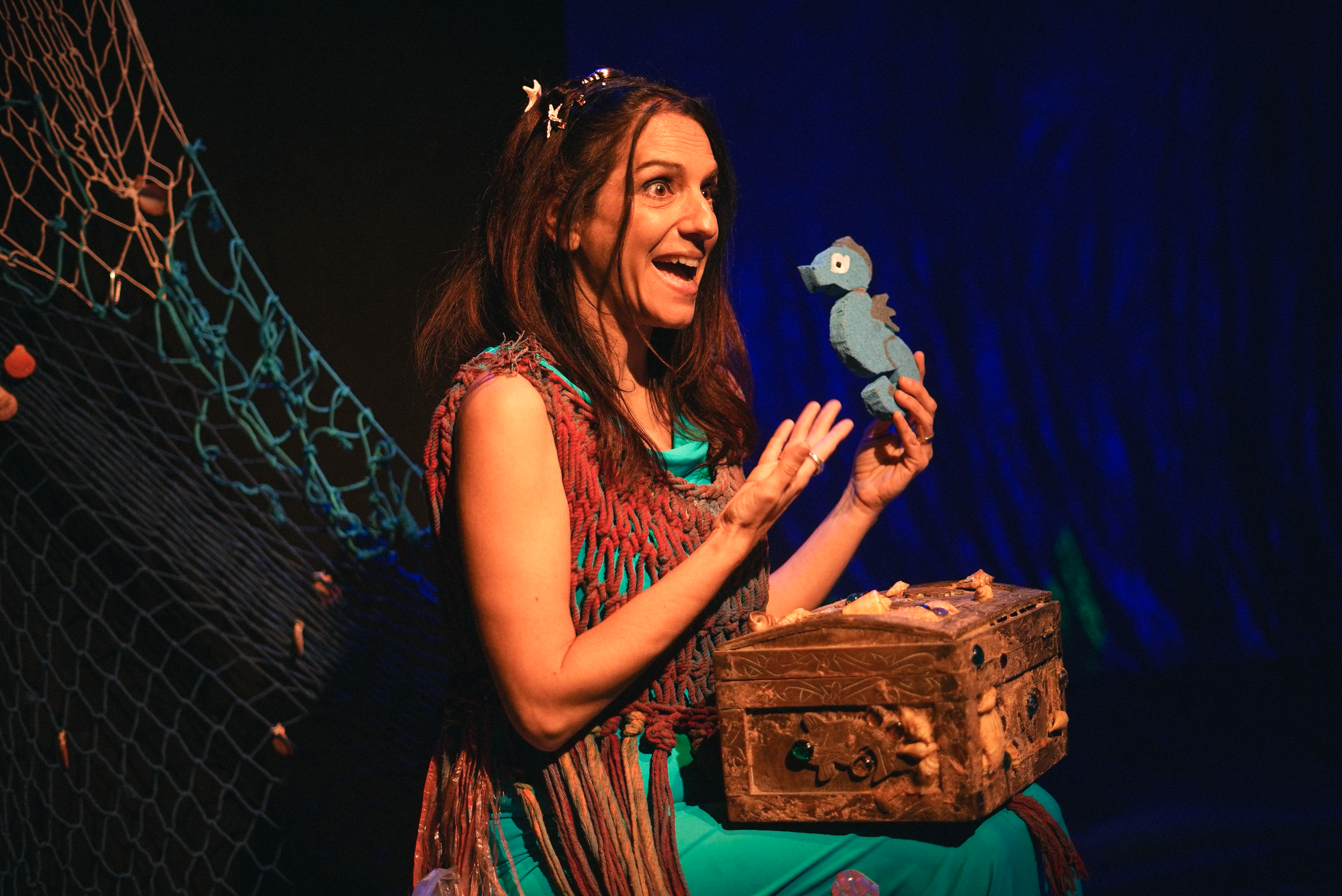 Una mujer con expresión sonriente sosteniendo un muñeco de peluche azul en un escenario con fondo oscuro y redes decorativas.
