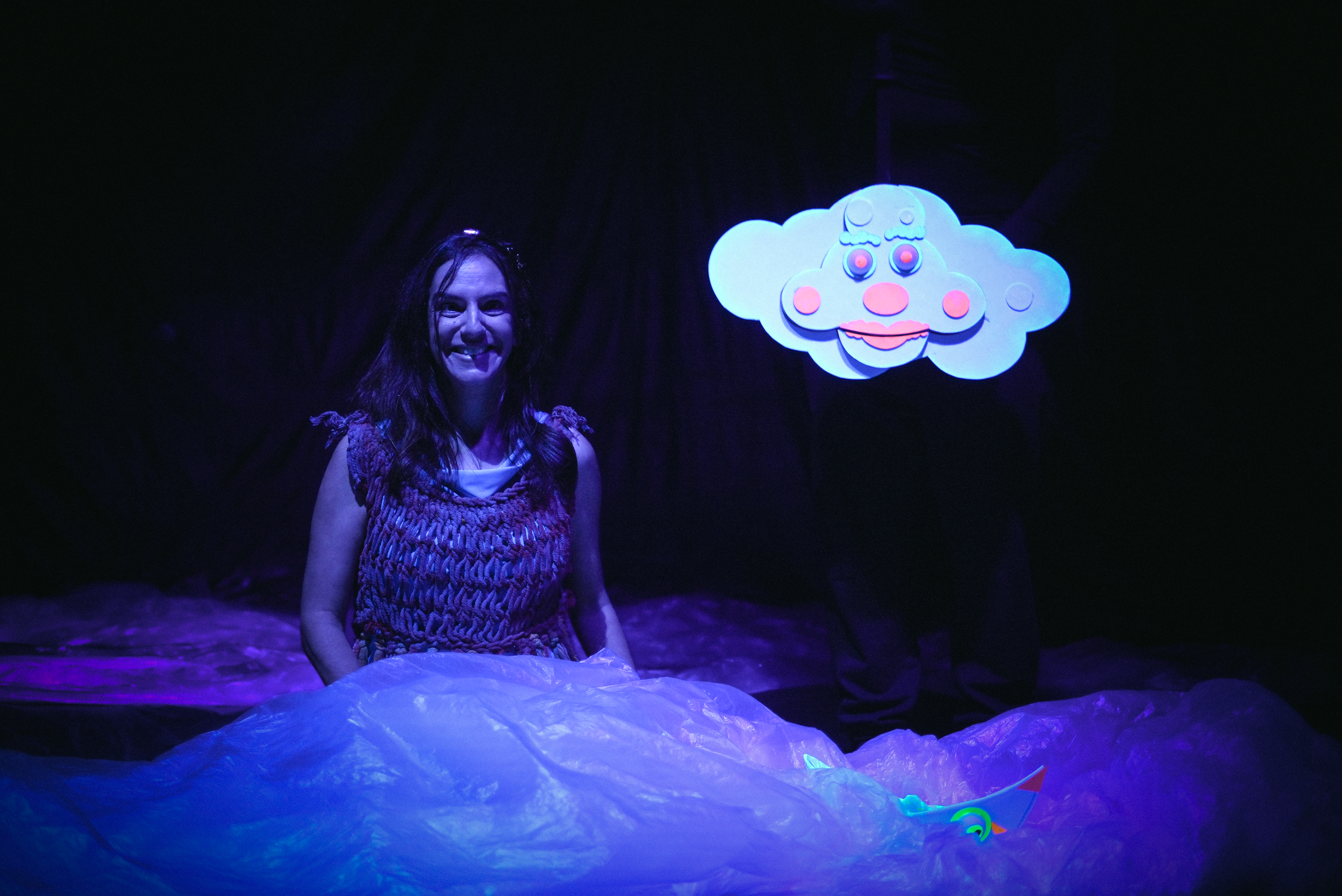 Una mujer sonriendo en un escenario oscuro con iluminación fluorescente, con una nube de felpa y un globo de dibujos animados de una cara de payaso encima.