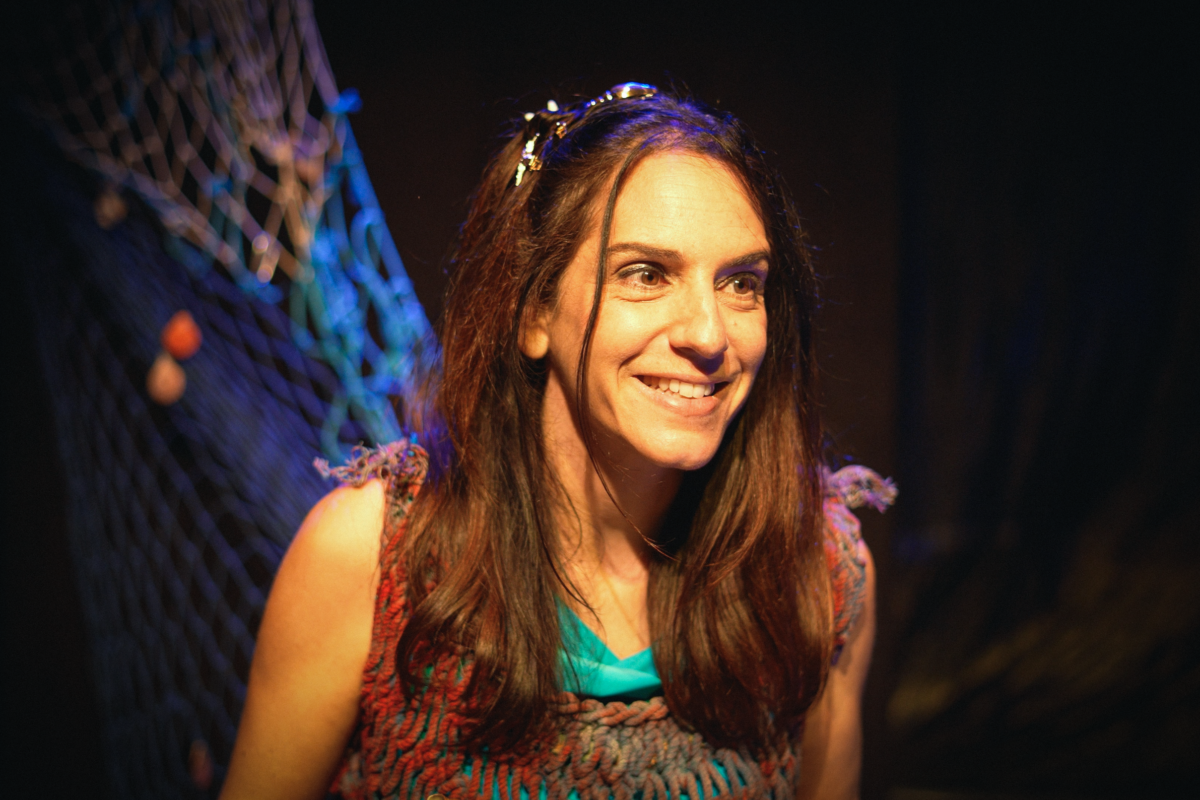 Mujer sonriendo con cabello rizado y camiseta de color azul, en un fondo oscuro con decoraciones de red y luces de colores.