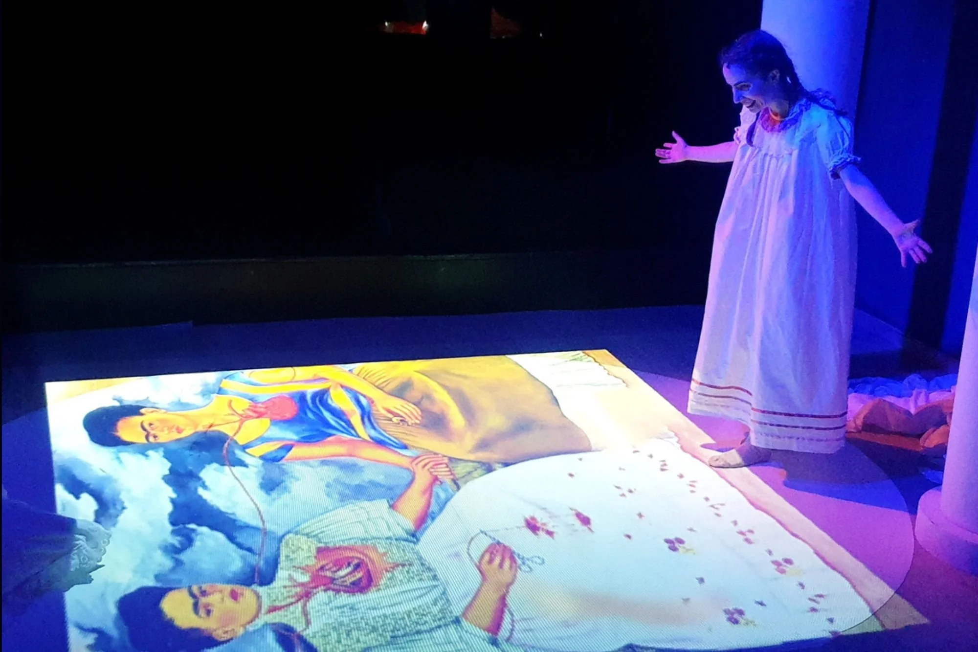 Niña en vestido blanco interactuando con un arte digital proyectado en el suelo que representa a una mujer en un cielo con nubes.