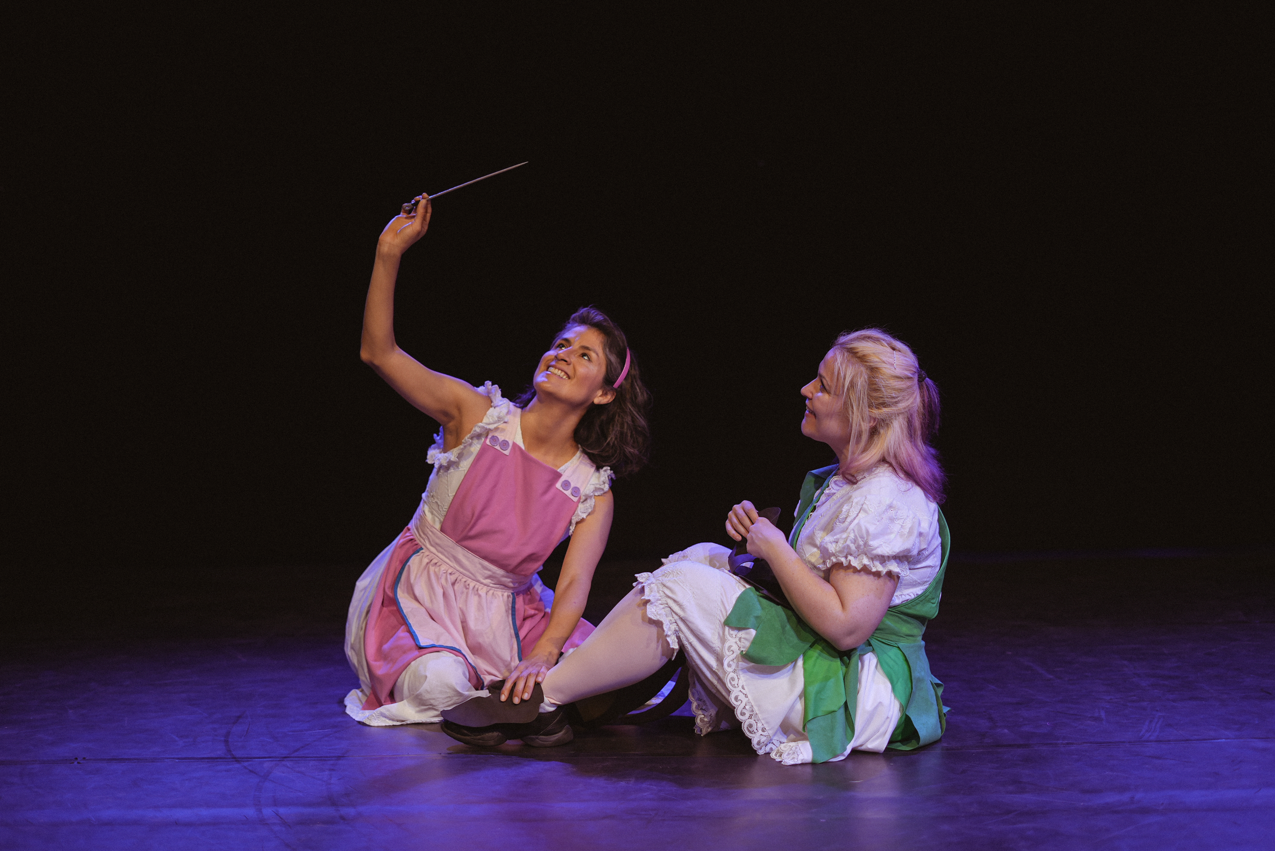 Dos mujeres en disfraces de teatro, una con un delantal rosa y otra con vestido verde y blanco, sentadas en el escenario con fondo negro, una de ellas sosteniendo un cuchillo de utilería y sonriendo.