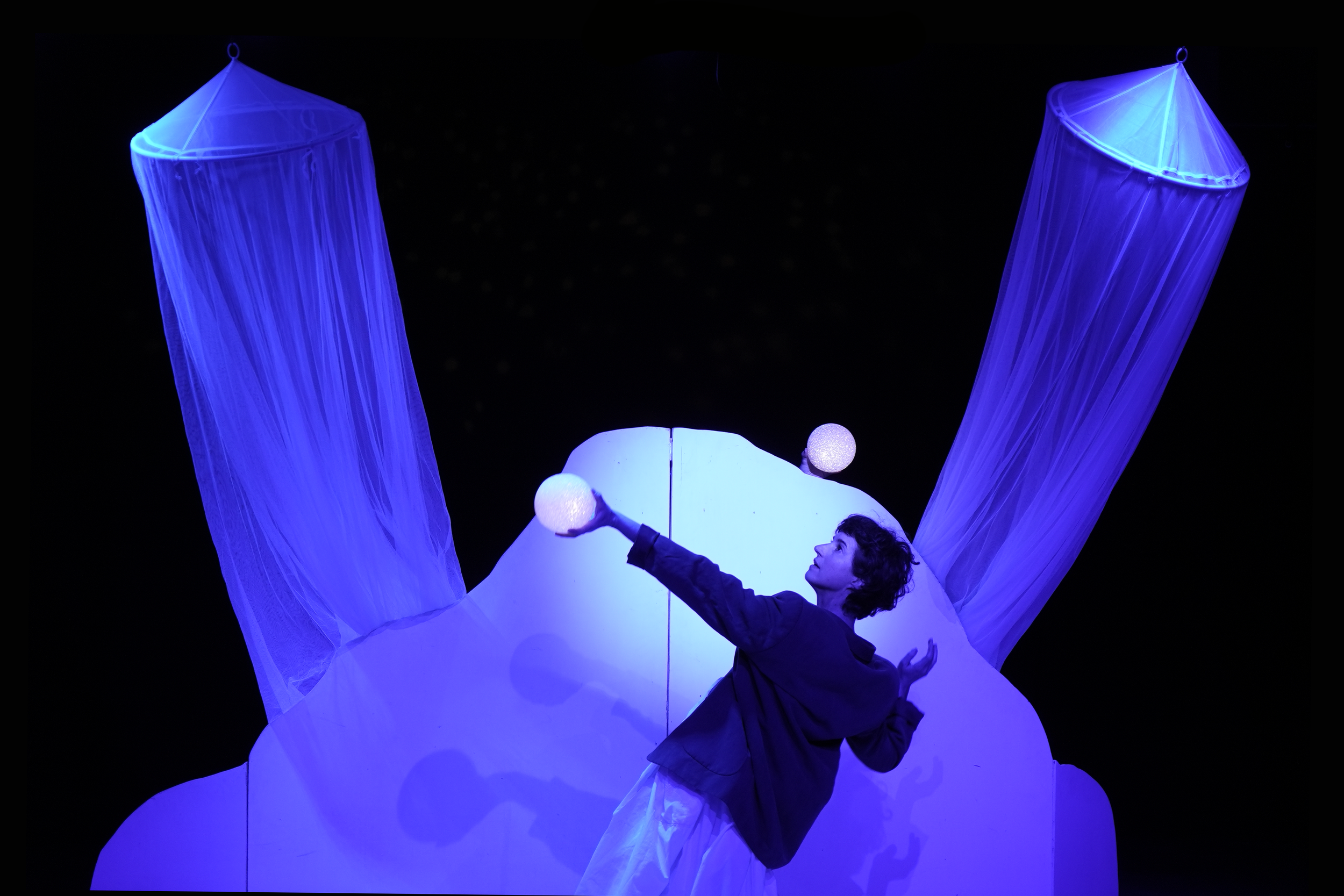 Persona actuando en un escenario con iluminación azul y decoraciones que parecen cortinas o telas, sosteniendo un globo en cada mano.