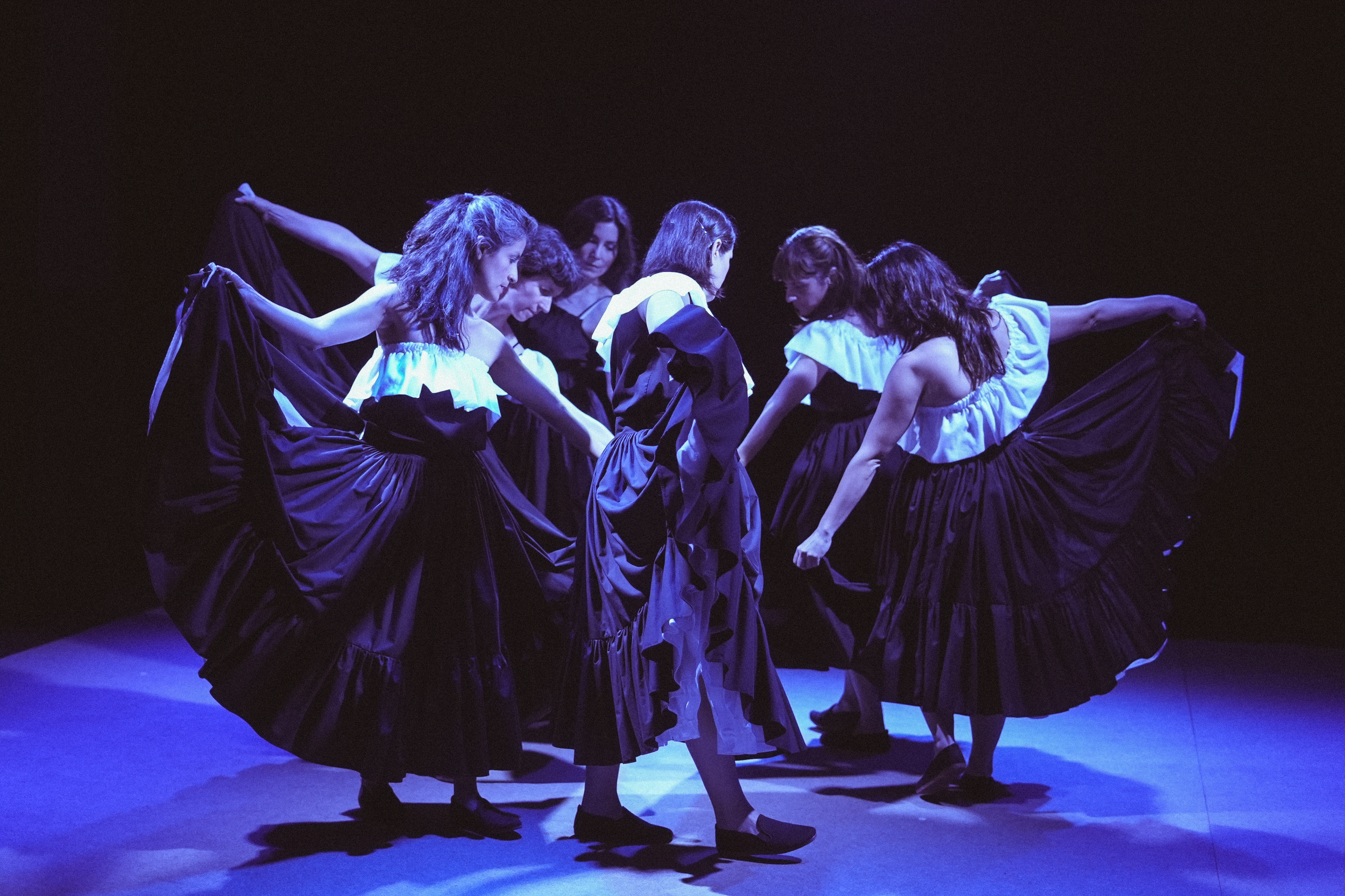 Grupo de bailarinas quemando en escenario oscuro, vestidas con trajes negros con mangas blancas, realizando una danza en círculo.
