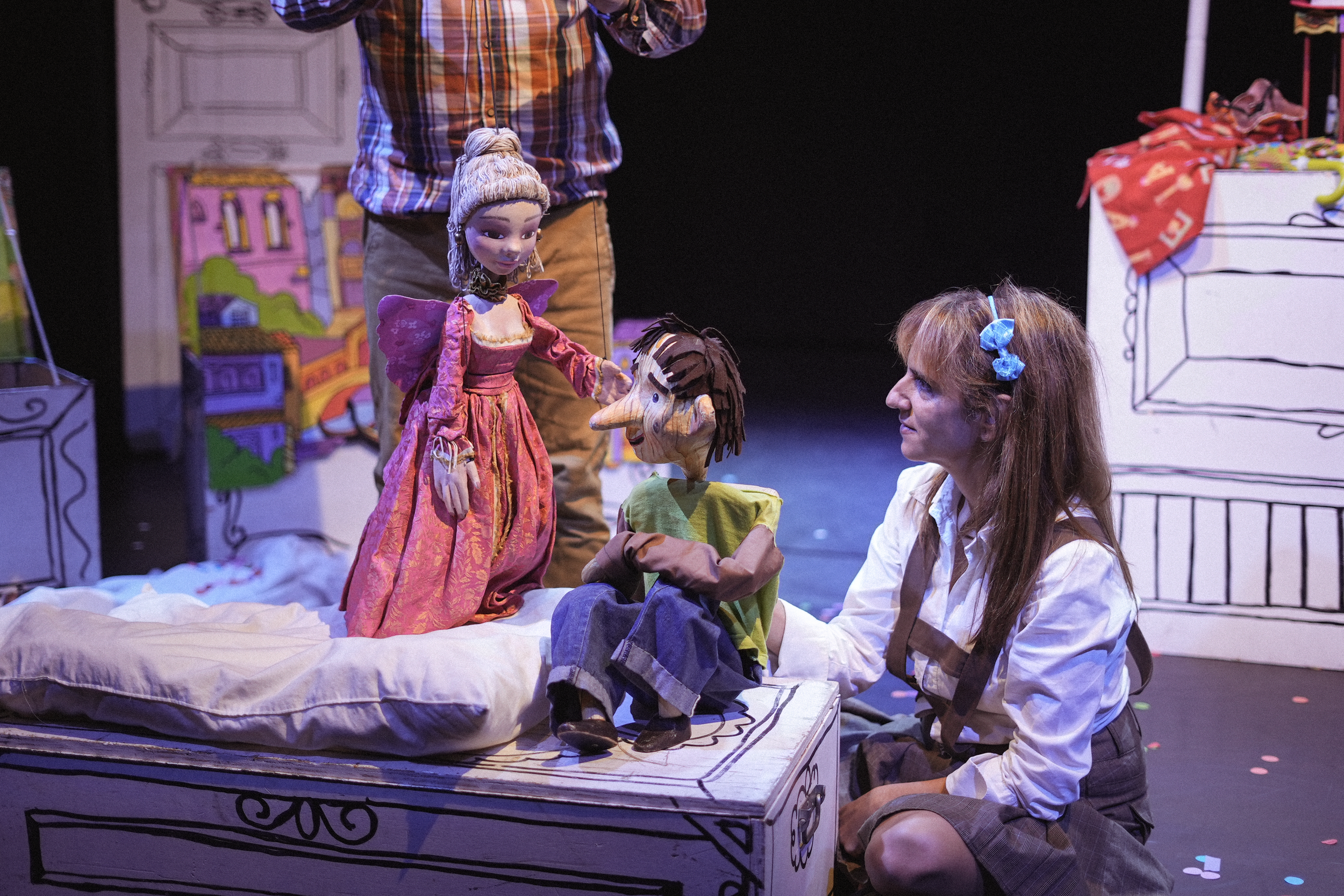 Técnicas de marionetas en un escenario teatral con personajes de marioneta y actores. Una mujer con cabello rizado y ropa vintage observa a una marioneta con cabello corto en un lugar decorado con dibujos de muebles y fondos de ciudad.