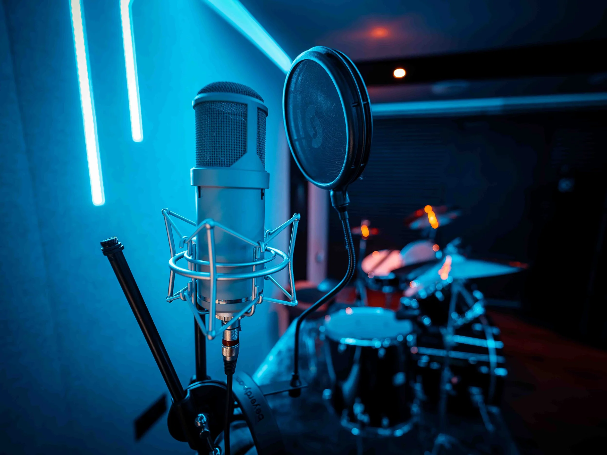 A studio microphone with a pop filter in front of it, illuminated by blue LED lights, with other musical equipment in the background.