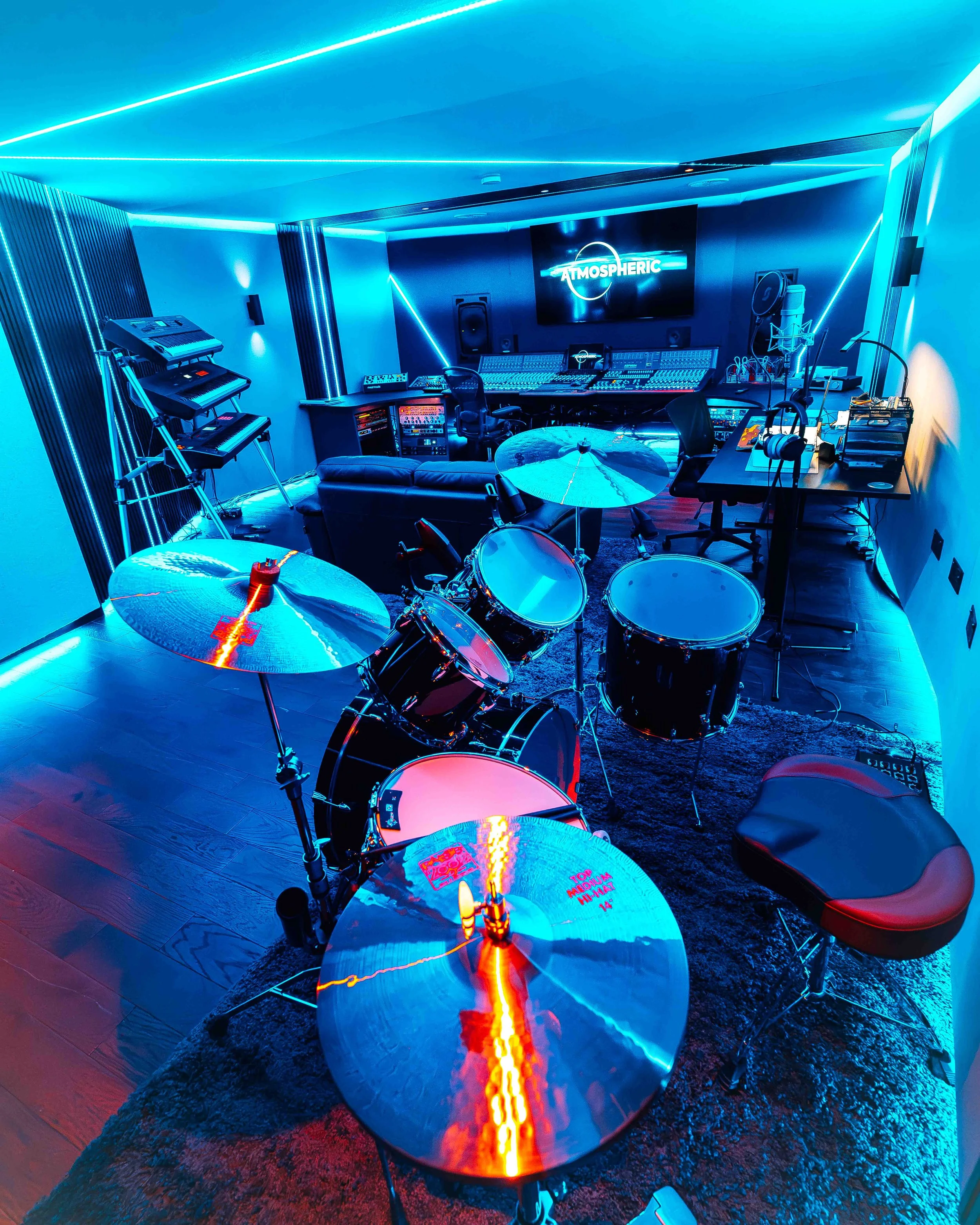 A music studio with blue neon lighting, featuring drums, keyboards, mixing console, speakers, headphones, and a large screen displaying the word 'ATMOSPHERIC'.