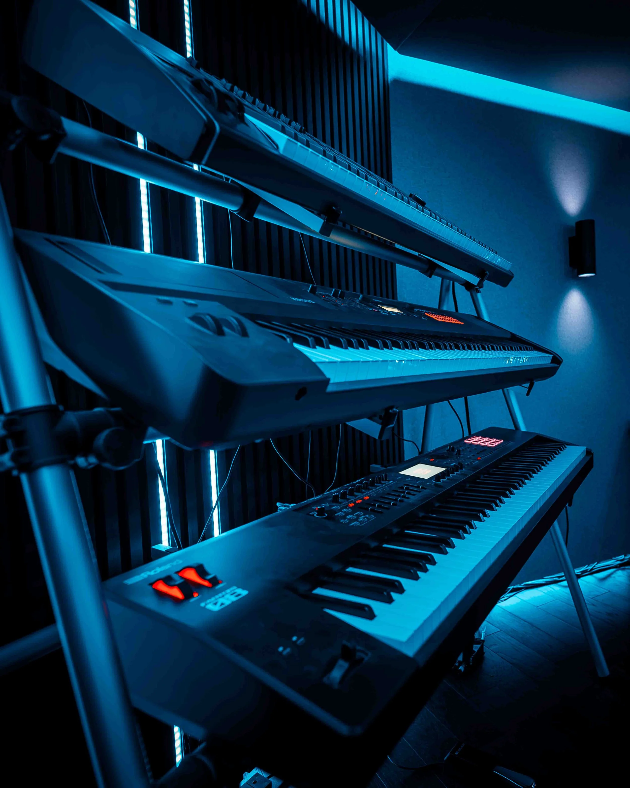 A digital music studio with multiple keyboards and synthesizers mounted on a metal rack in a dark room illuminated by blue LED lighting.