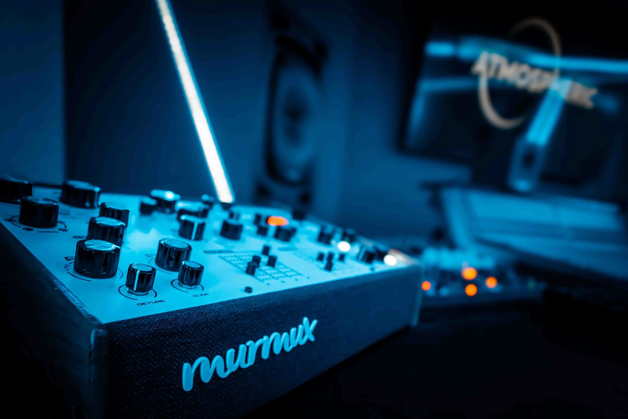 Close-up of a Murmux Synth with illuminated blue lighting, featuring knobs and sliders, with a blurred background of audio equipment.