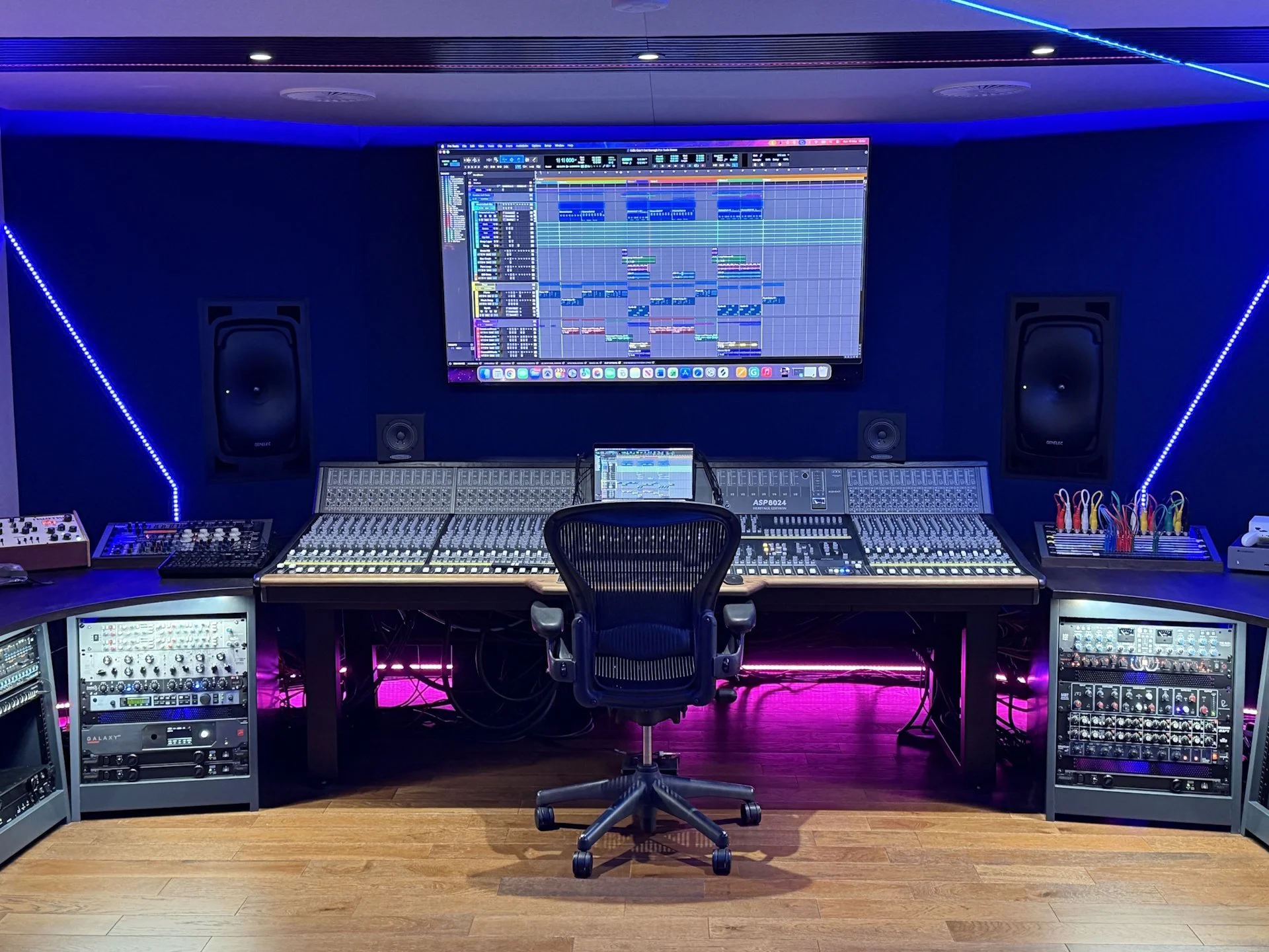 A professional music recording studio with a large mixing console, speakers, computer monitors displaying music production software, and vibrant blue LED accent lighting.