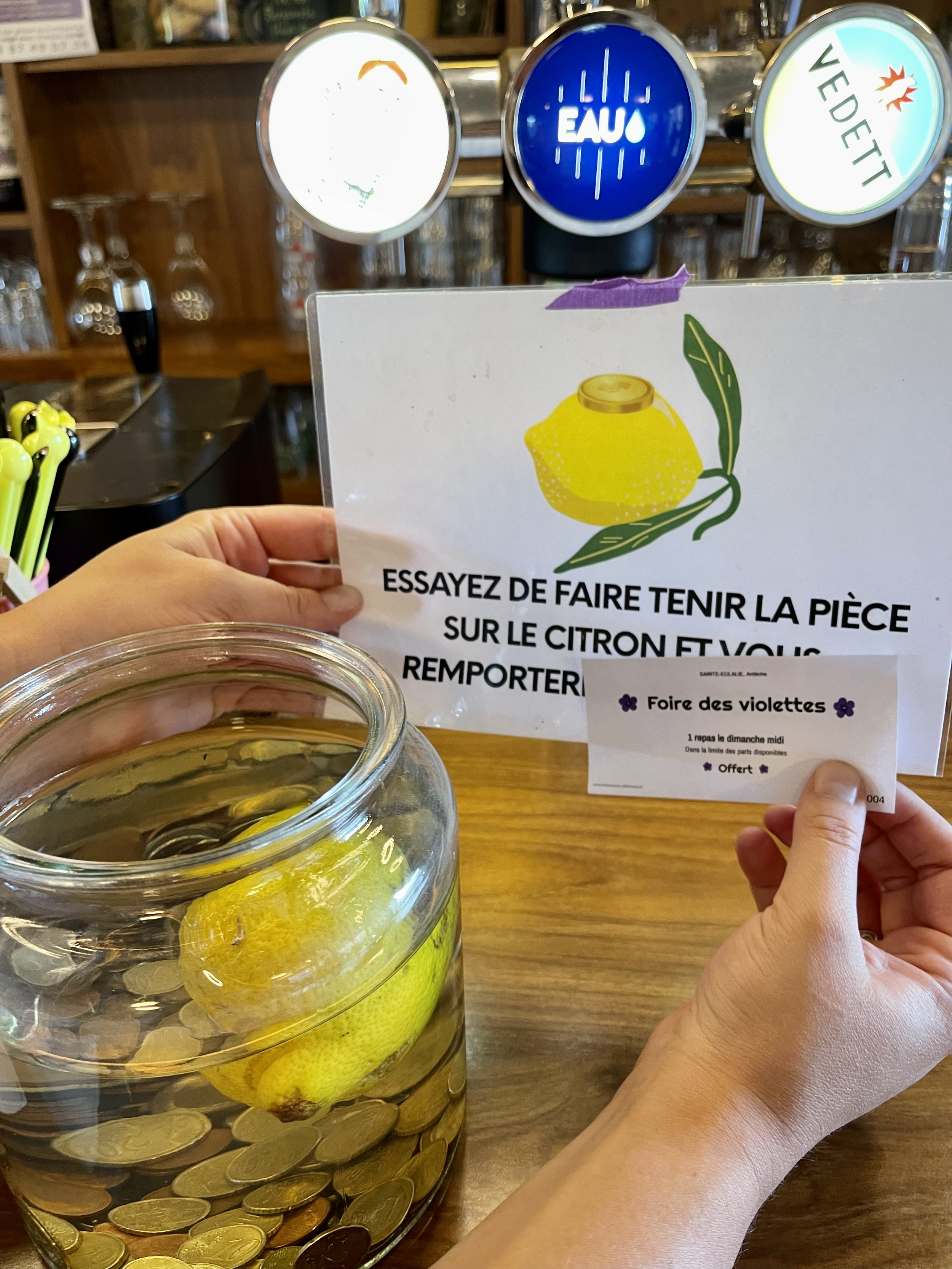 Un grand bocal rempli de pièces de monnaie avec un citron à l'intérieur, placé sur un comptoir en bois. En arrière-plan, un panneau avec une illustration de citron et du texte en français, ainsi que des inscriptions sur les robinets de boisson.