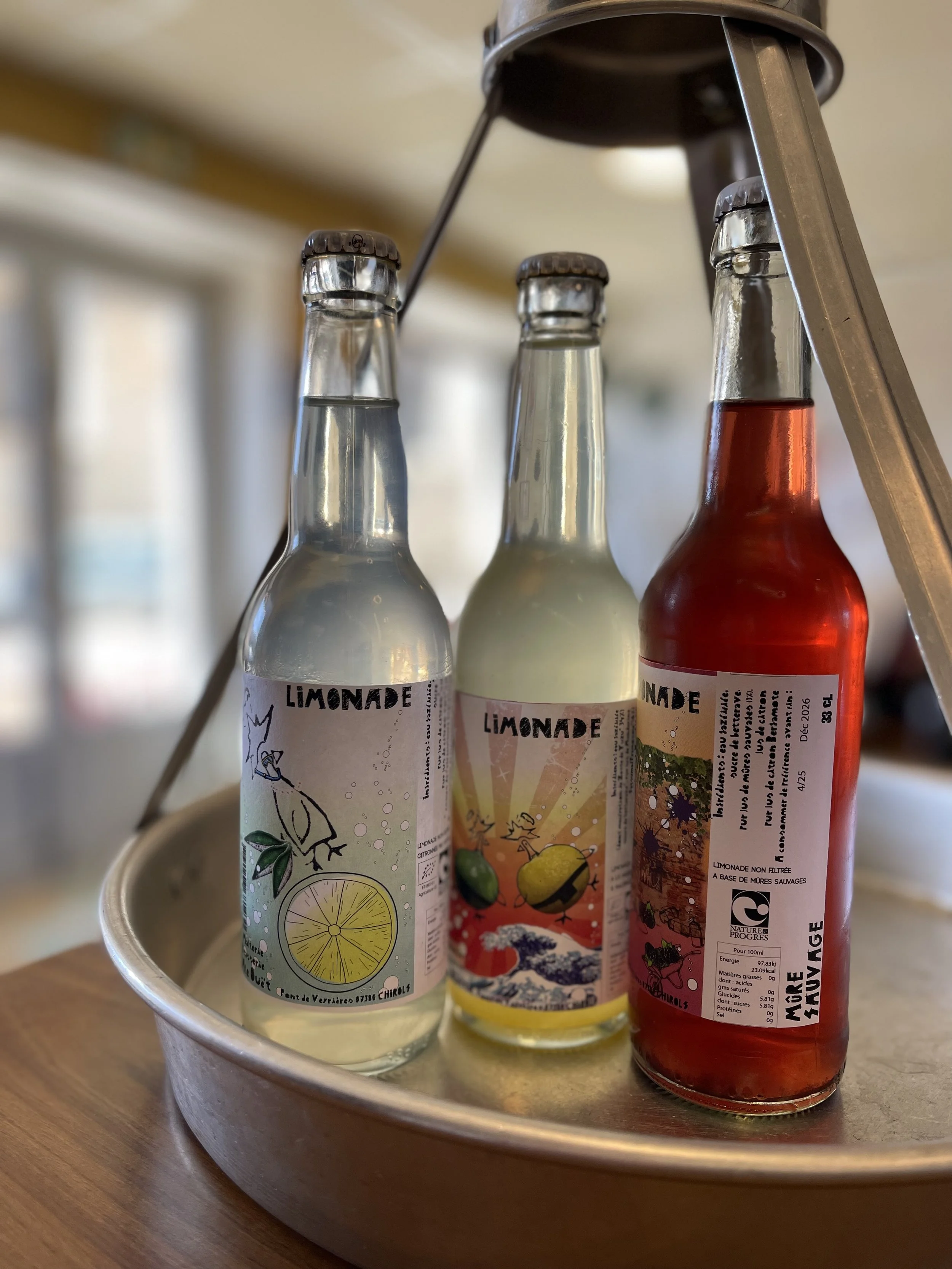 Trois bouteilles de limonade aux étiquettes colorées, disposées sur un plateau en métal, avec une échelle en arrière-plan.