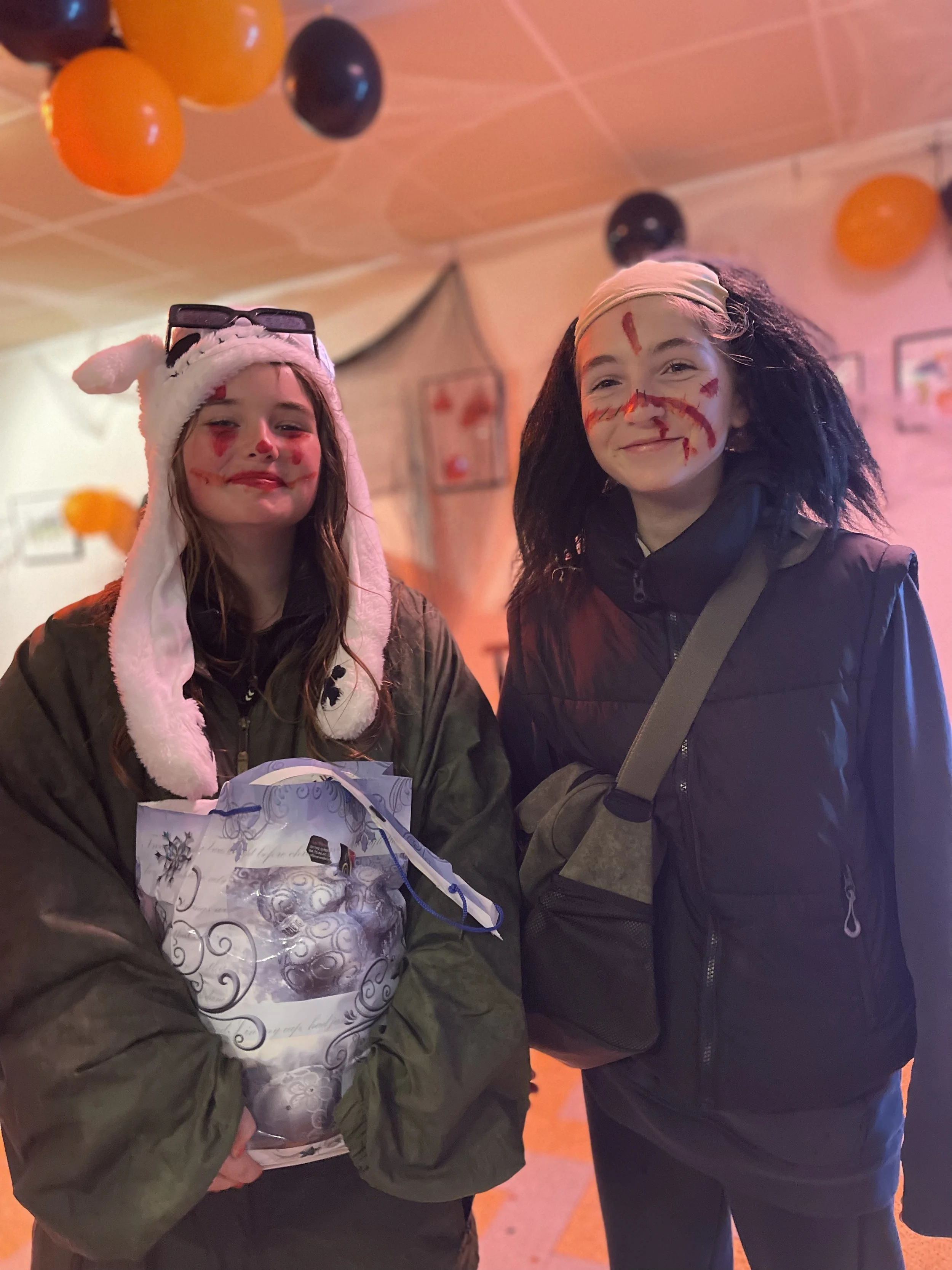 Deux jeunes filles souriantes déguisées pour Halloween avec du maquillage effrayant, portant des vêtements chauds, se tenant dans une pièce décorée avec des ballons orange et noirs.