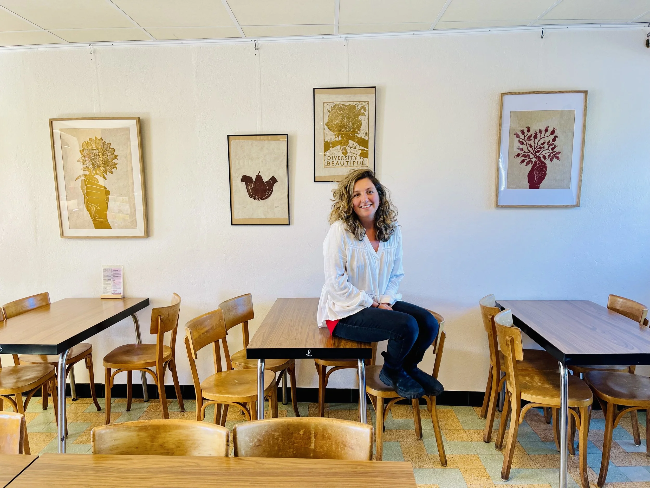 Une femme assise sur une table dans un café, entourée de chaises en bois, décorée de plusieurs œuvres d'art sur le mur.