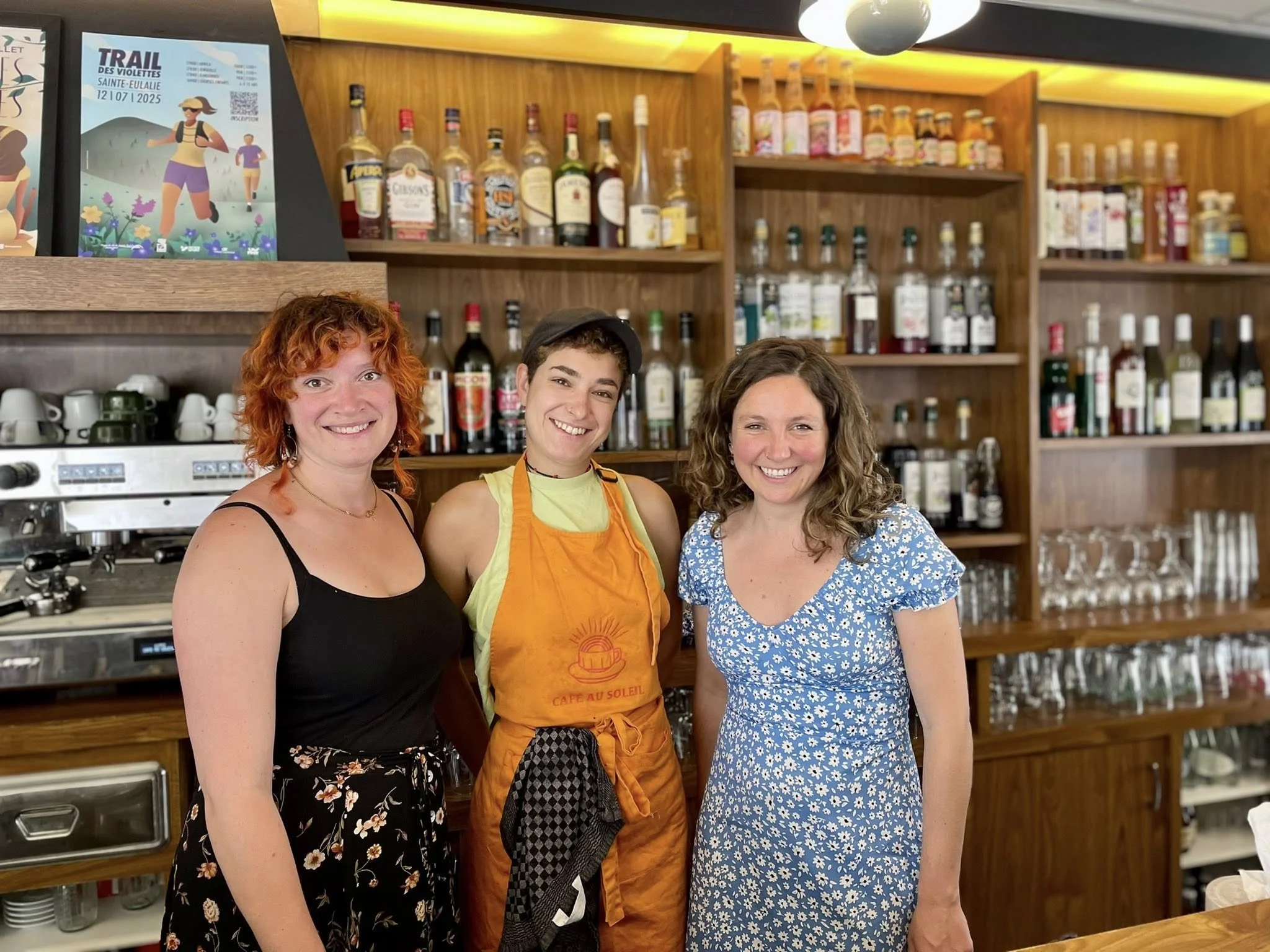 Trois femmes souriantes posent derrière le comptoir d'un bar ou café, avec des étagères de bouteilles d'alcool et de verres en arrière-plan.