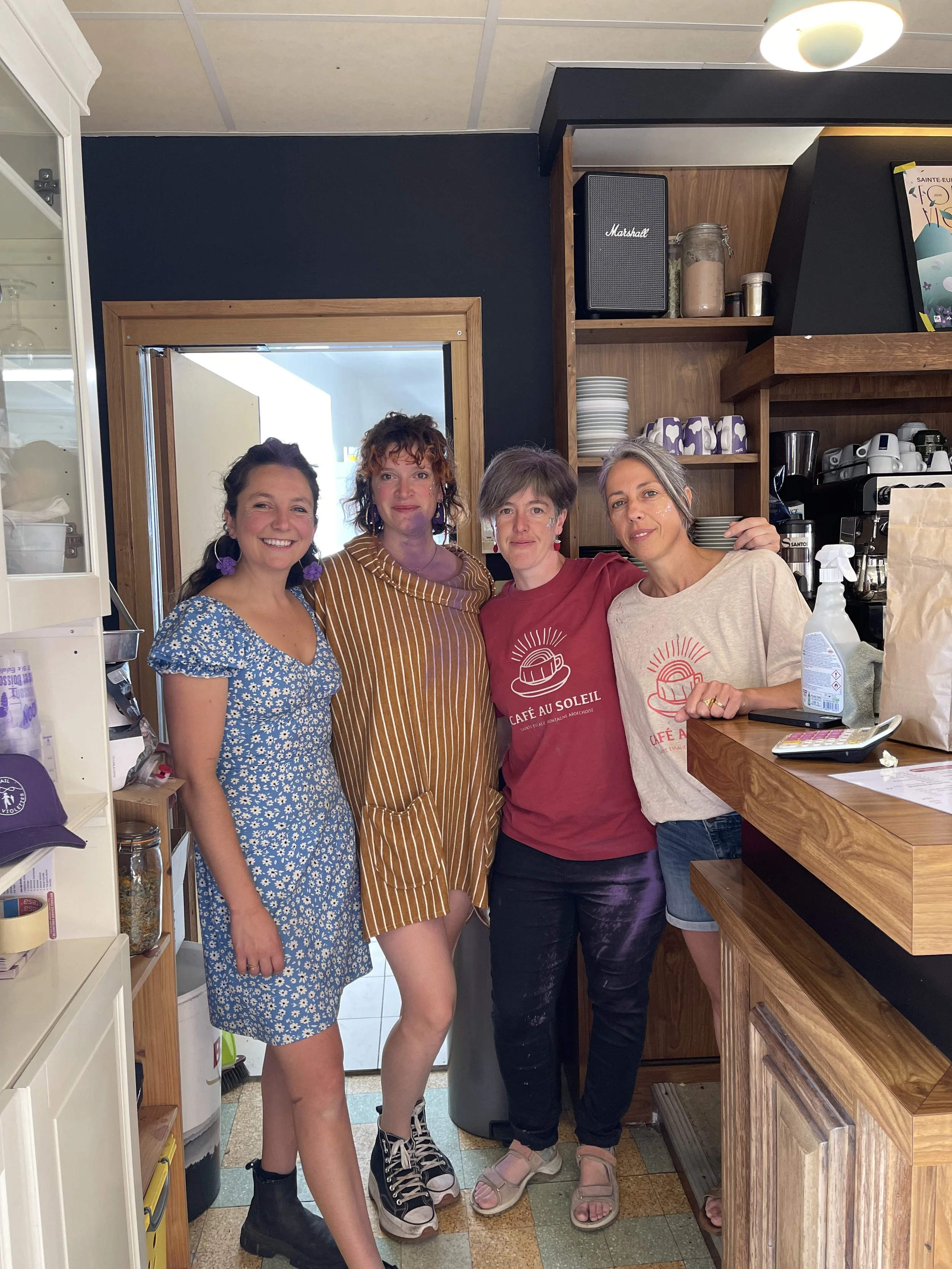 Quatre femmes posant ensemble dans un café, derrière le comptoir en bois, avec des étagères contenant des tasses, assiettes et appareils photo, en arrière-plan, dans un espace intérieur avec murs en bois.