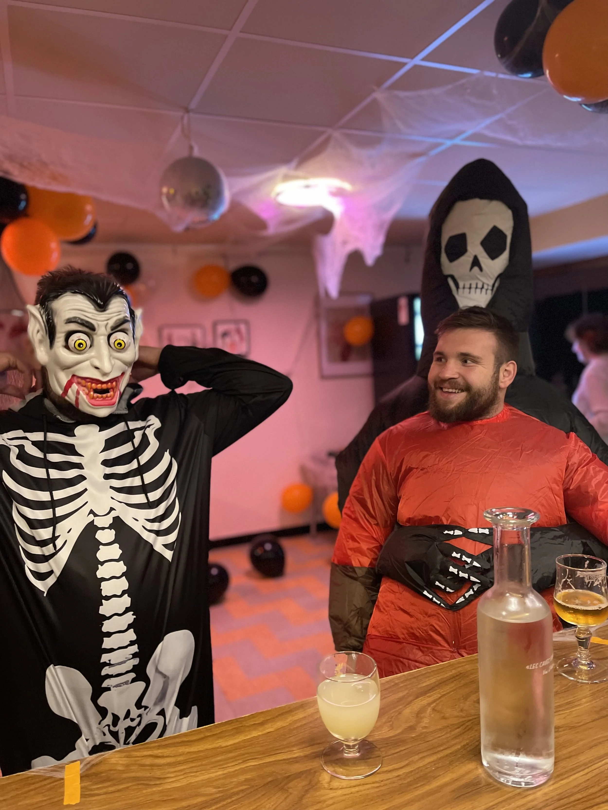 Une fête d'Halloween avec des personnes déguisées en squelette et en mort-vivant, décorée avec des ballons orange et noirs, des toiles d'araignées et un symbolique faux cercueil au fond.