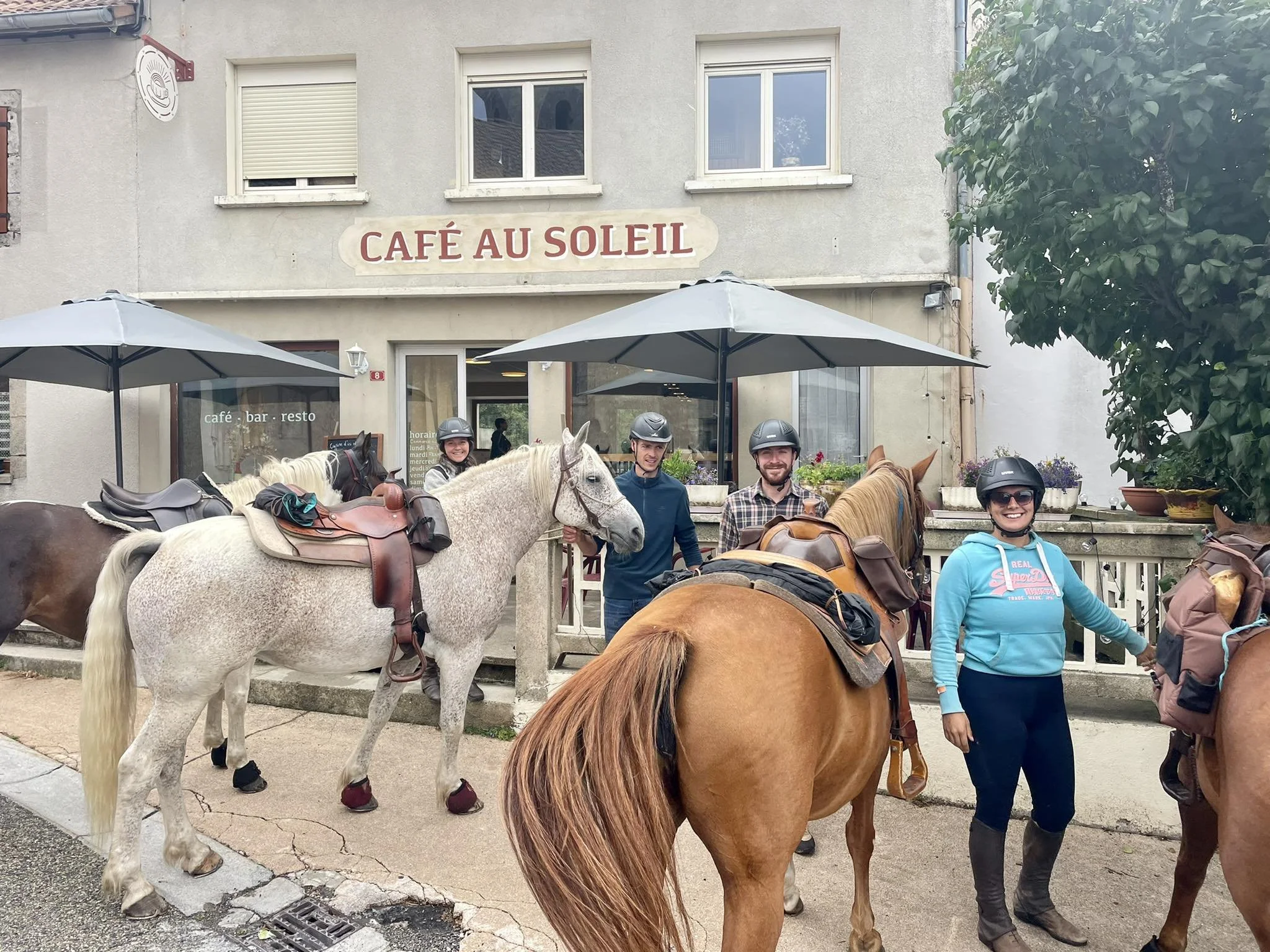Groupe de personnes à cheval devant un café appelé 'Café au Soleil', portant des casques, avec deux chevaux blancs et un cheval brun avec des selles et des sacs. Le café a une enseigne et des parasols en face.