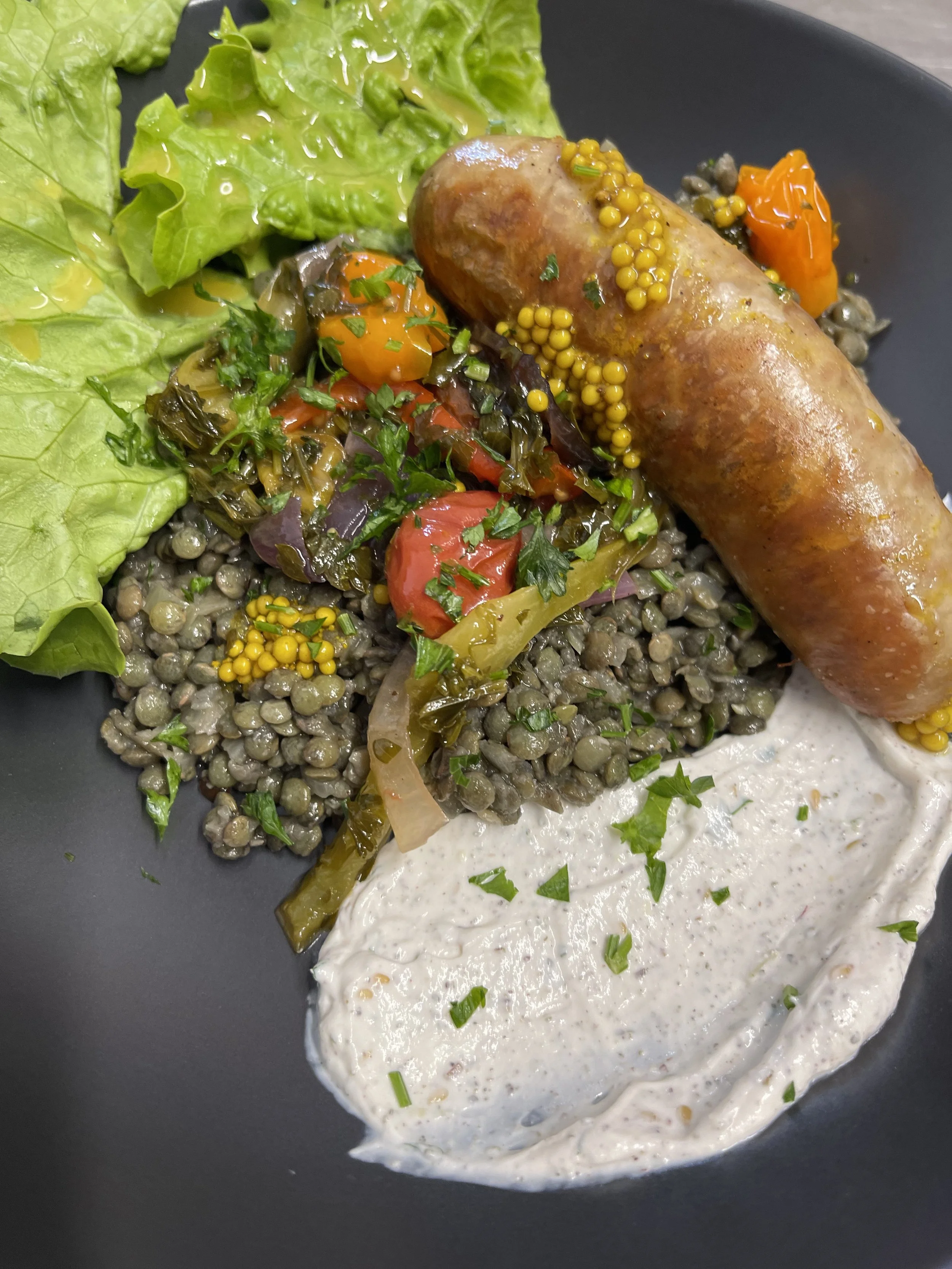 Assiette de nourriture avec une saucisse, un mélange de légumes sautés, une salade verte et une sauce blanche garnie de persil.