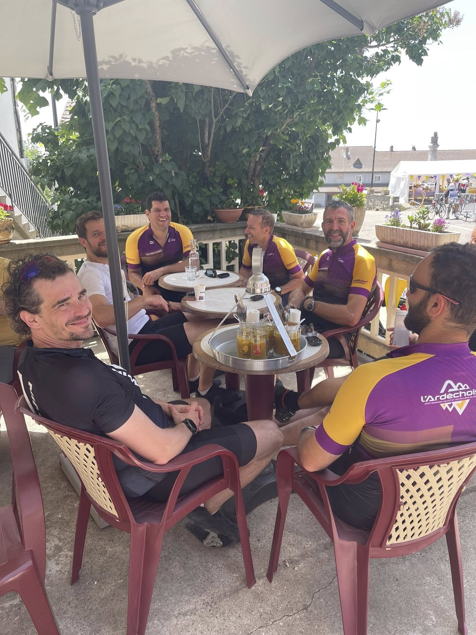 Groupe de huit personnes assises autour d'une table en terrasse, certains portant des maillots de cyclisme violets et jaunes, en train de partager un moment de convivialité. Plantes en pot et parasol en toile blanche en arrière-plan.