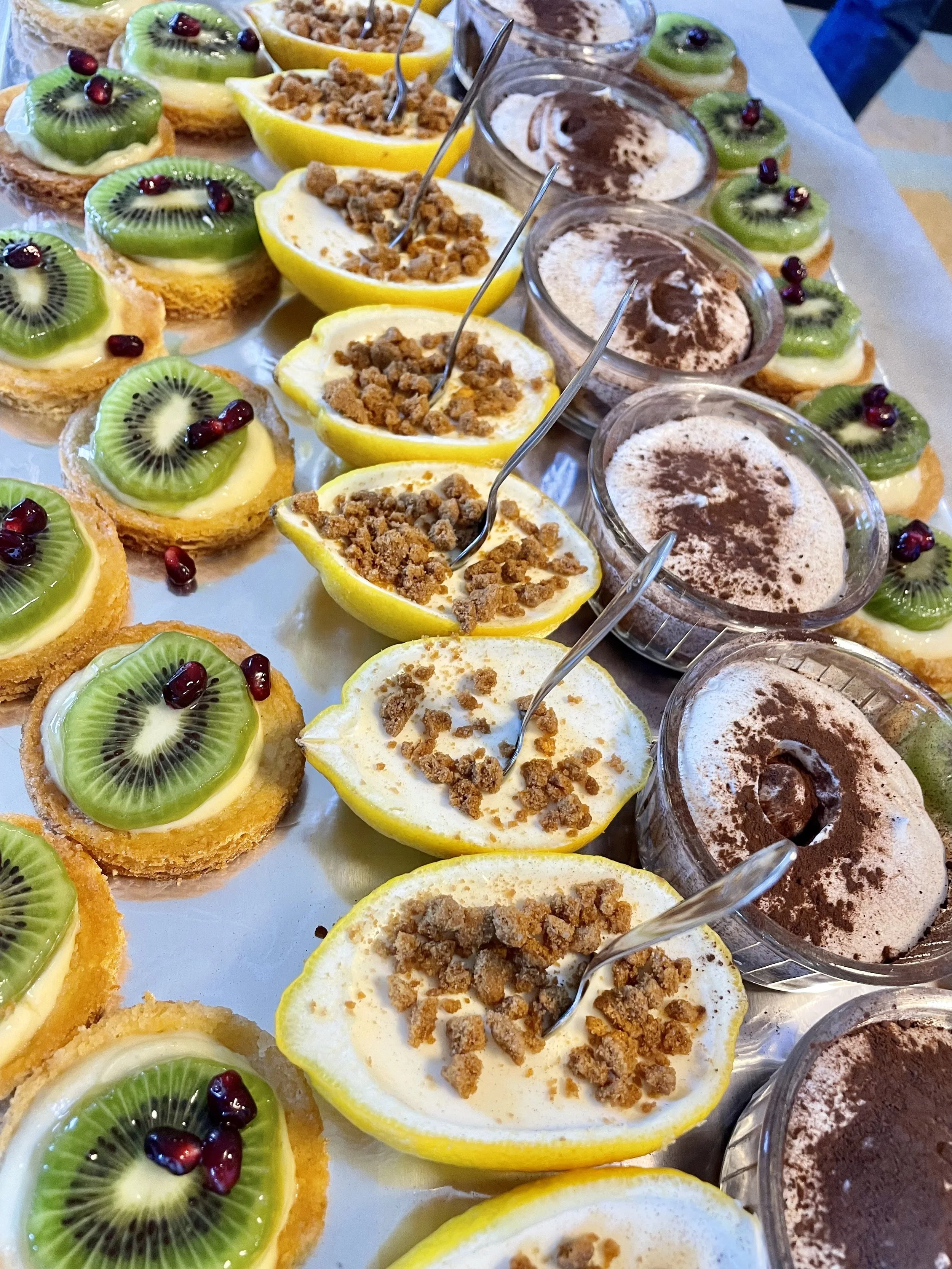 Assortiment de desserts en portions individuelles : tartes aux kiwis, citrons aux crumble, et crèmes au chocolat, garnies de fruits et de poudre de cacao.