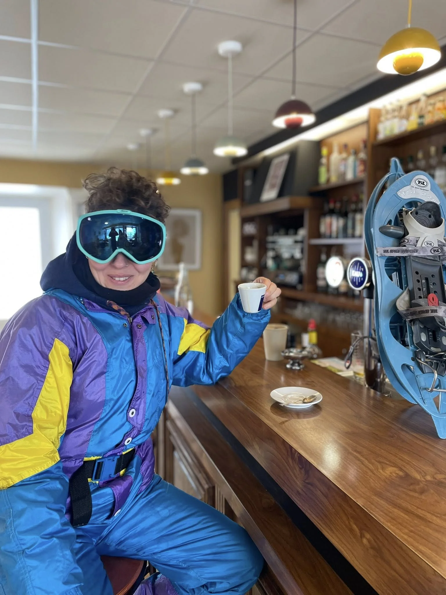 Personne en tenue de ski colorée assise à un comptoir de café, portant des lunettes de ski, tenant une tasse, dans un café ou un chalet.