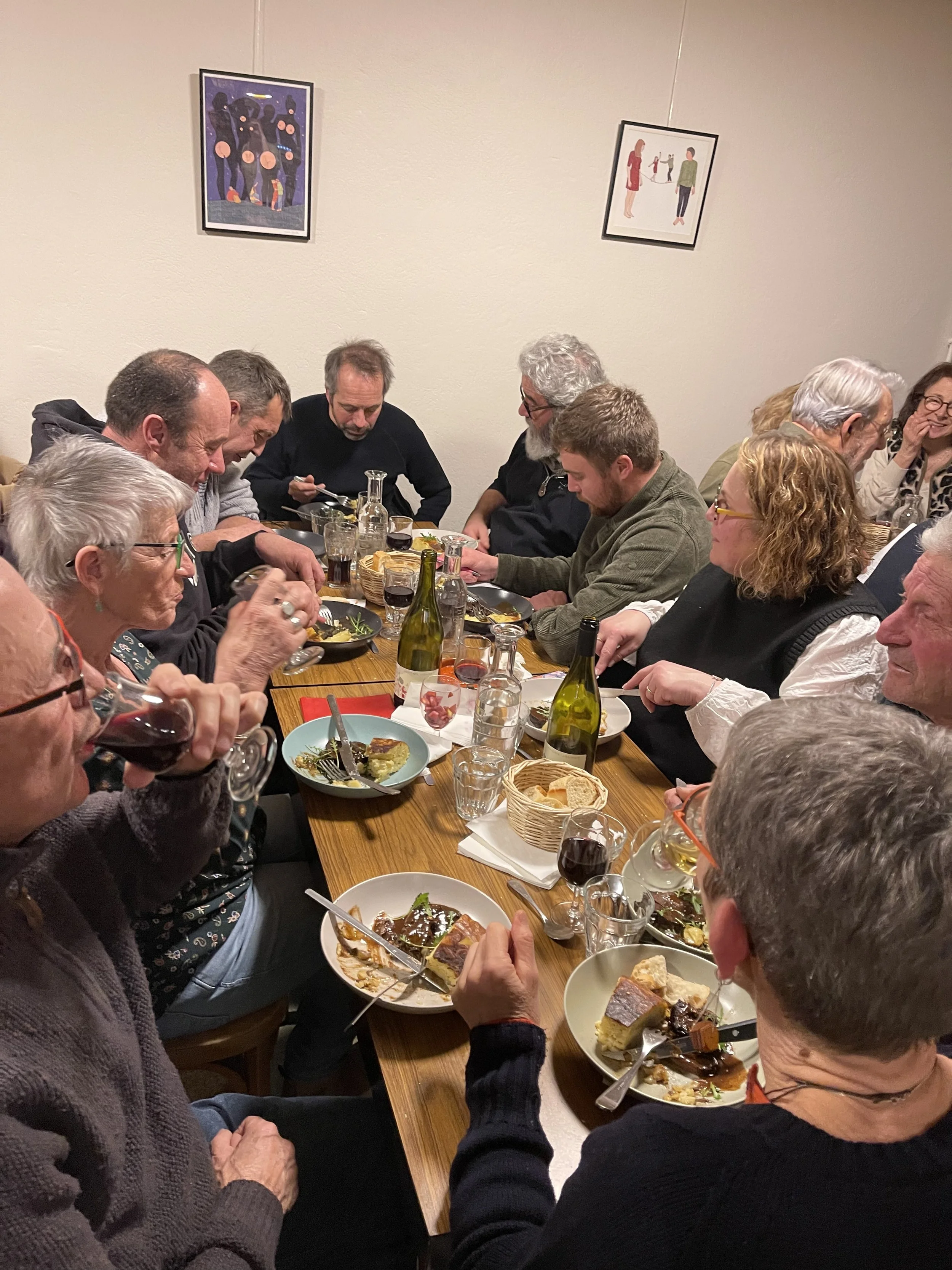 Groupe de personnes lors d'un repas convivial autour d'une longue table, avec des assiettes de nourriture, des verres de vin et des bouteilles, dans une pièce décorée avec deux œuvres d'art au mur.