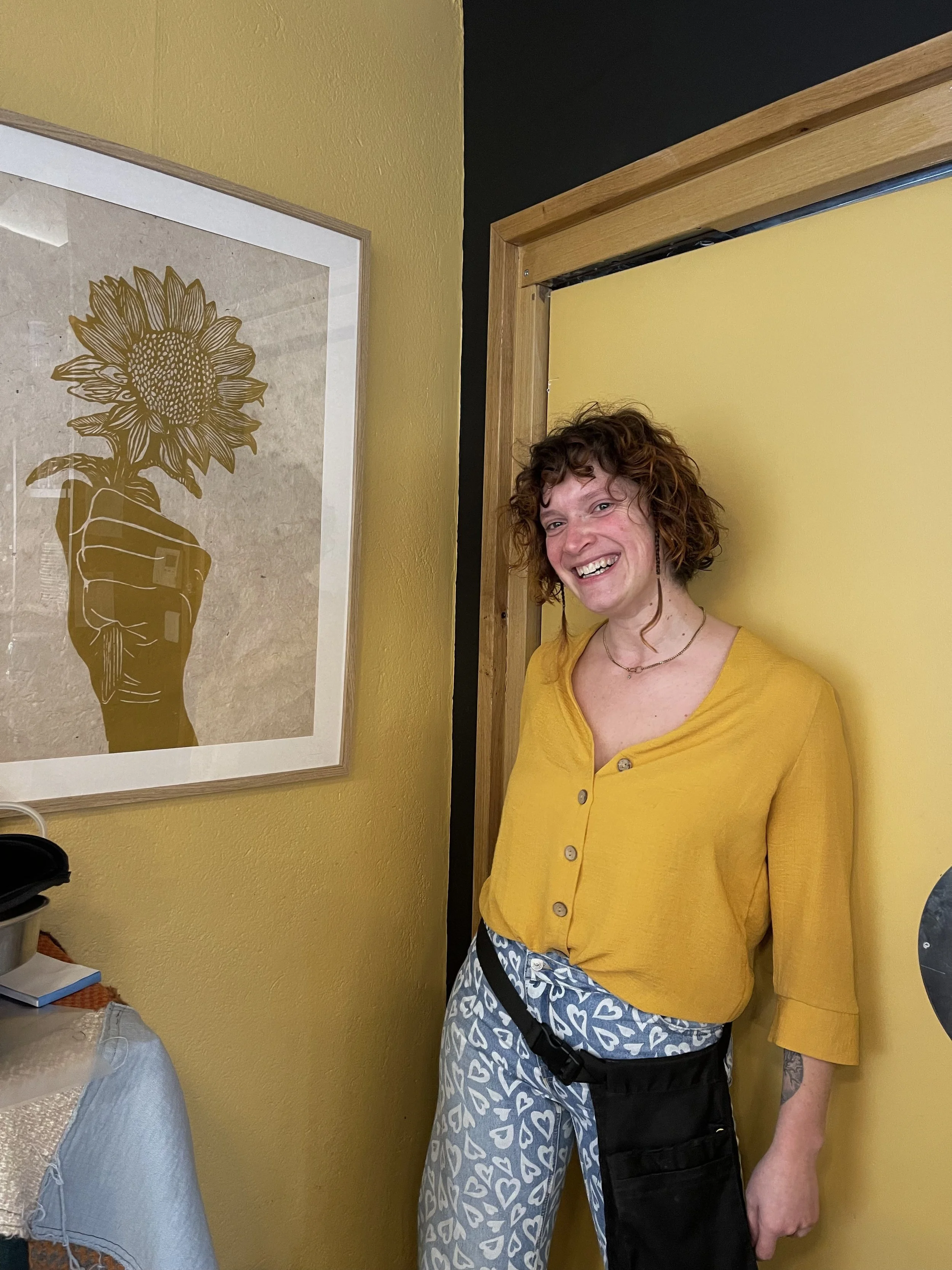 Une femme souriante portant un chemisier jaune et un pantalon à motifs de cœurs, debout à côté d'une porte jaune dans une pièce avec un tableau représentant une main tenant un tournesol.