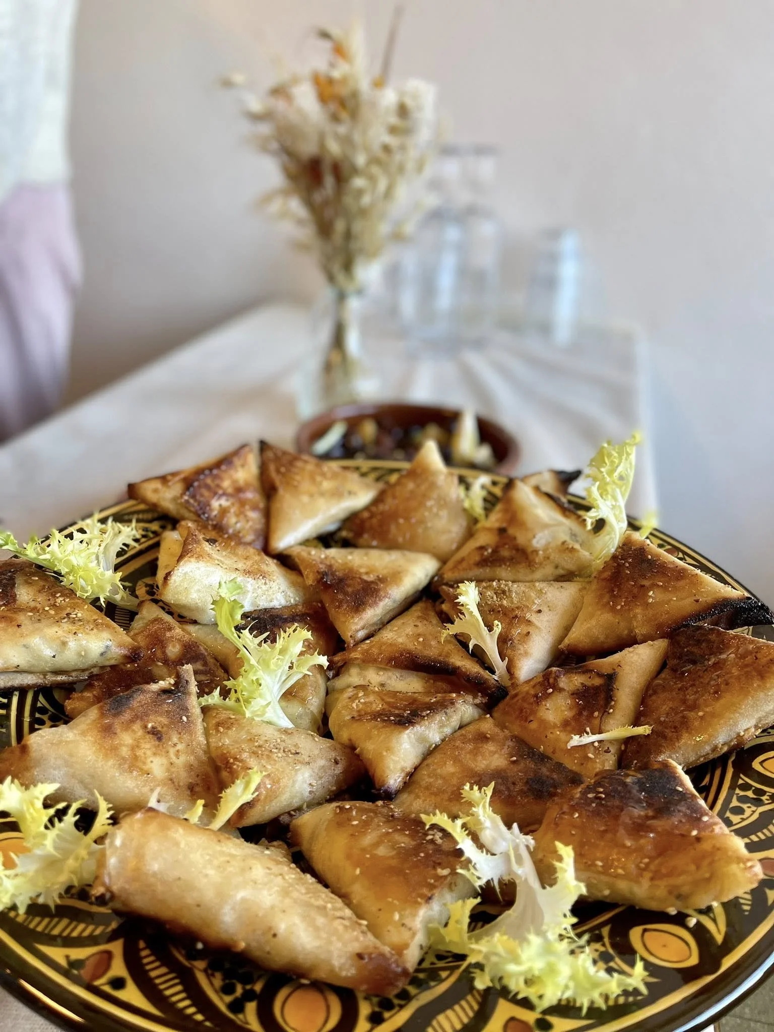 Assiette de samoussas dorés avec de la salade verte, sur une table avec un vase de fleurs séchées et des verres en arrière-plan.