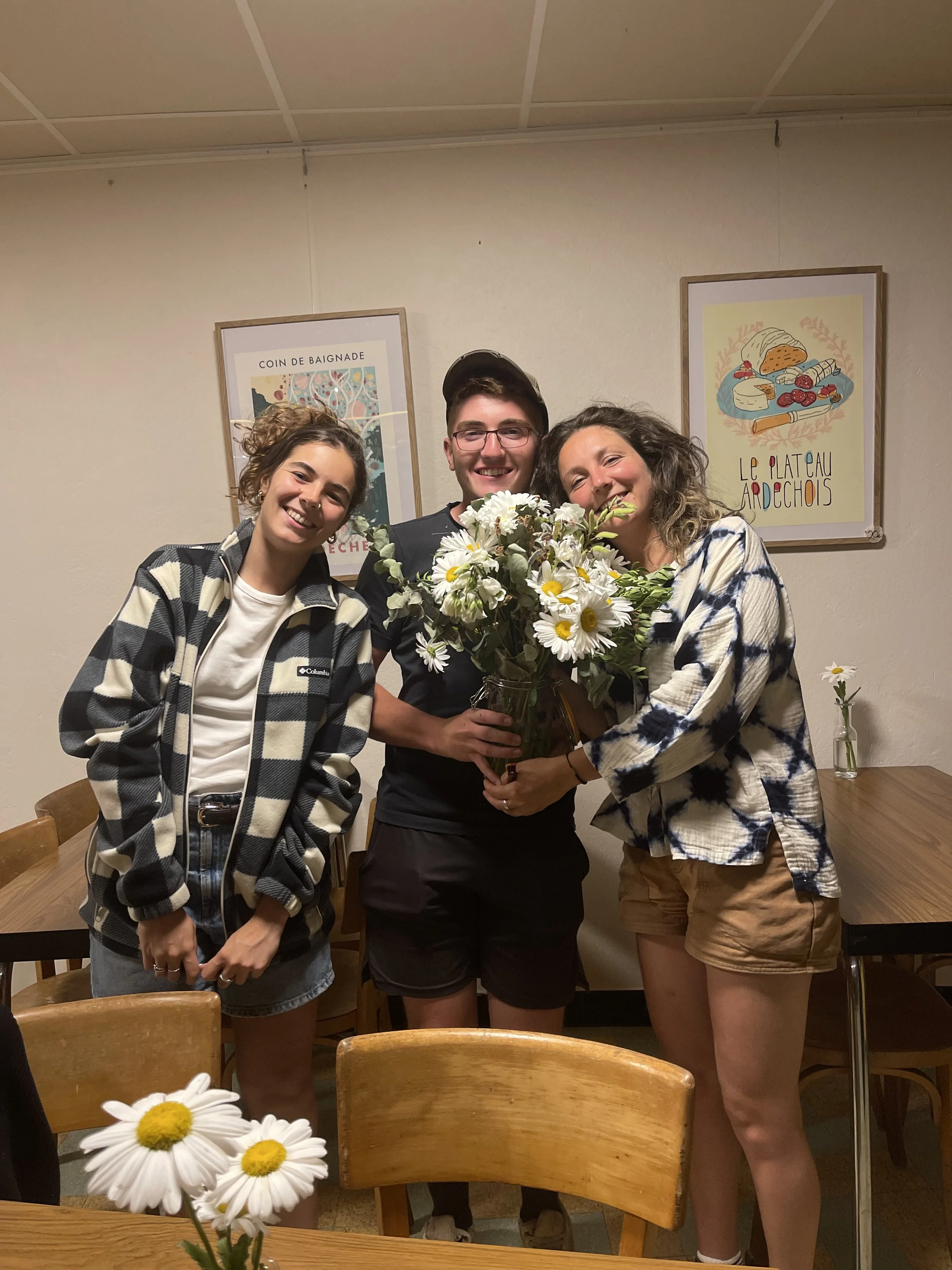 Trois personnes souriantes posent avec un bouquet de fleurs, dans une salle à manger. Deux femmes à gauche et à droite, une jeune homme au centre. La salle a des affiches avec du texte en français sur le mur.
