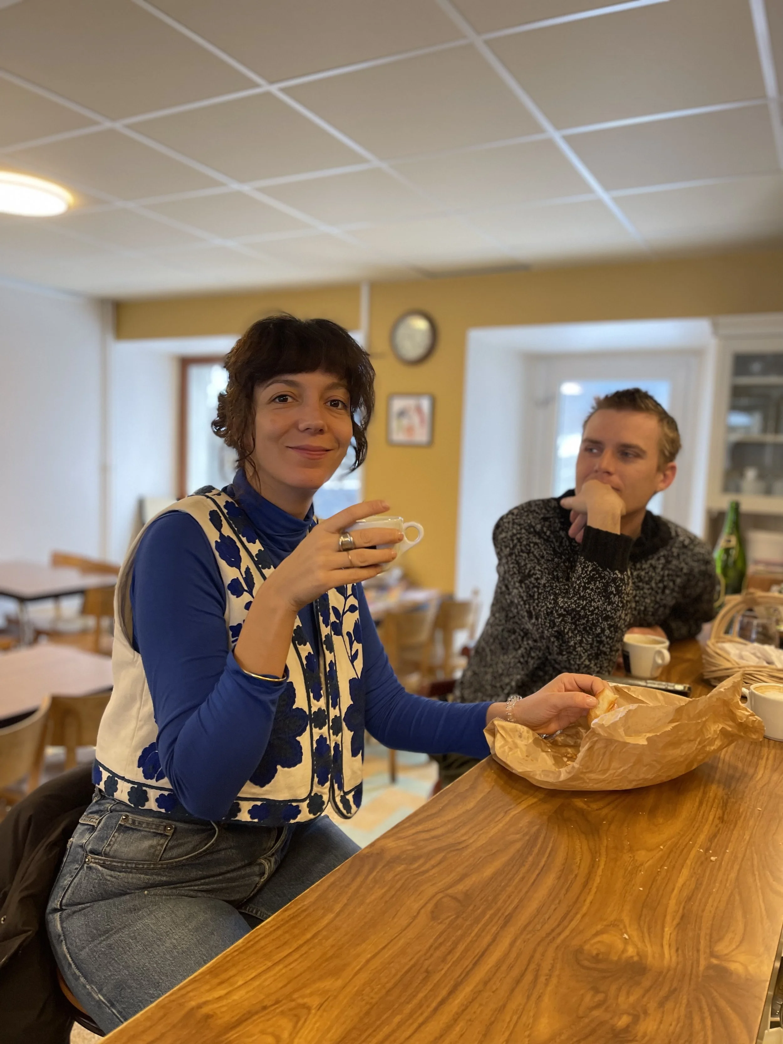 Une femme souriante tenant une tasse de café, assise à un bar en bois, avec un homme assis à côté en arrière-plan dans un café ou restaurant.