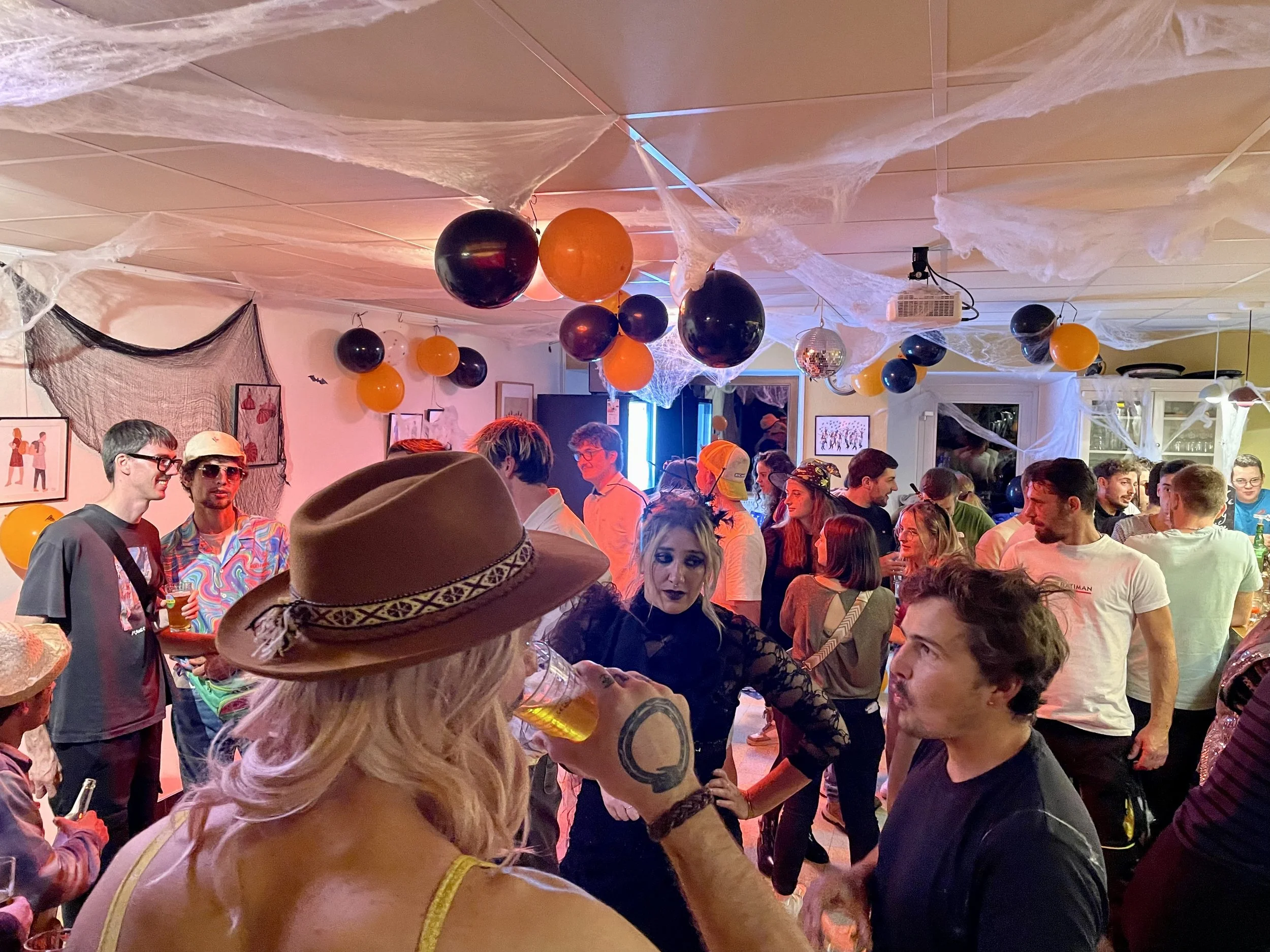 Fête d'Halloween avec décor de toiles d'araignées, ballons orange et noirs, et un groupe de personnes qui socialisent dans une pièce.