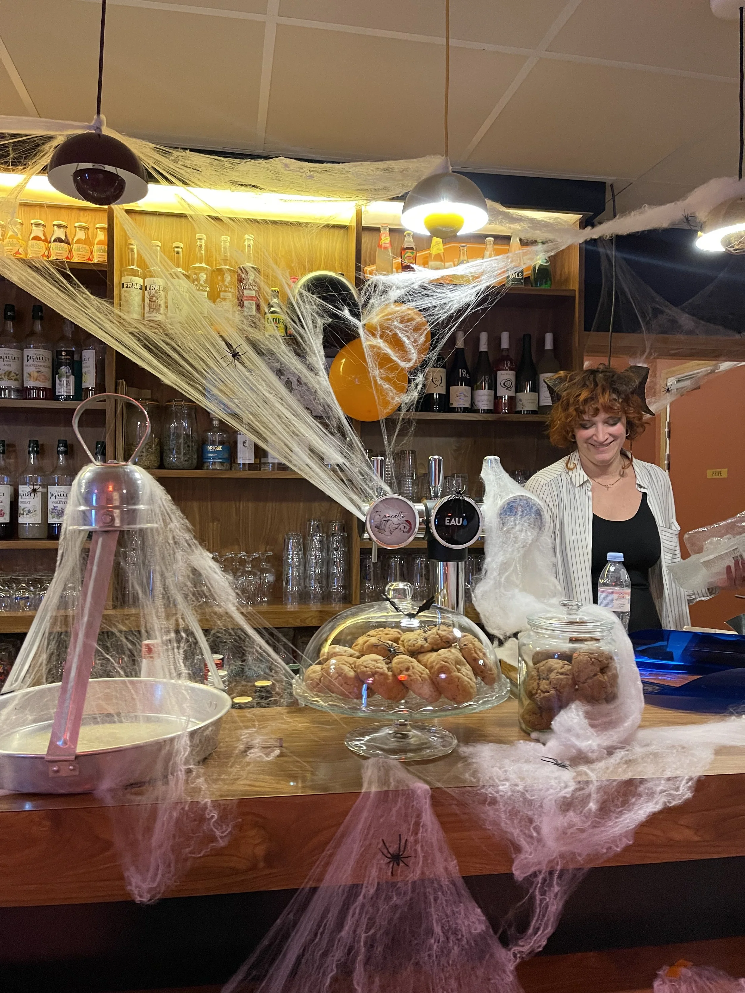 Une femme souriante derrière un bar décoré avec des toiles d'araignée en plastique, des araignées en plastique, et des ballons orange et noir. Sur le bar, il y a des cookies sous un cloche en verre, des bouteilles de boissons, et des accessoires de d