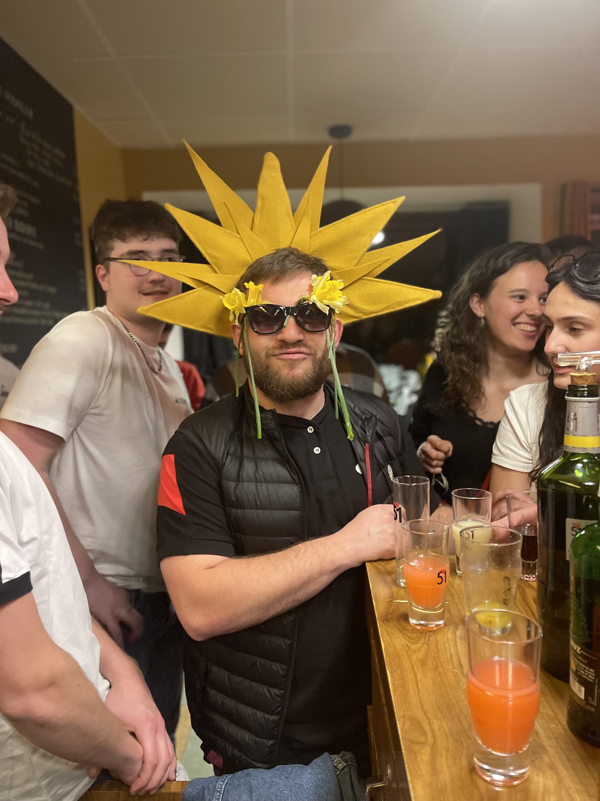 Groupe de personnes dans un bar, homme au centre portant un chapeau en forme de soleil jaune, avec de grandes lunettes noires et des fleurs jaunes, tenant une boisson. D'autres personnes autour qui sourient et discutent.