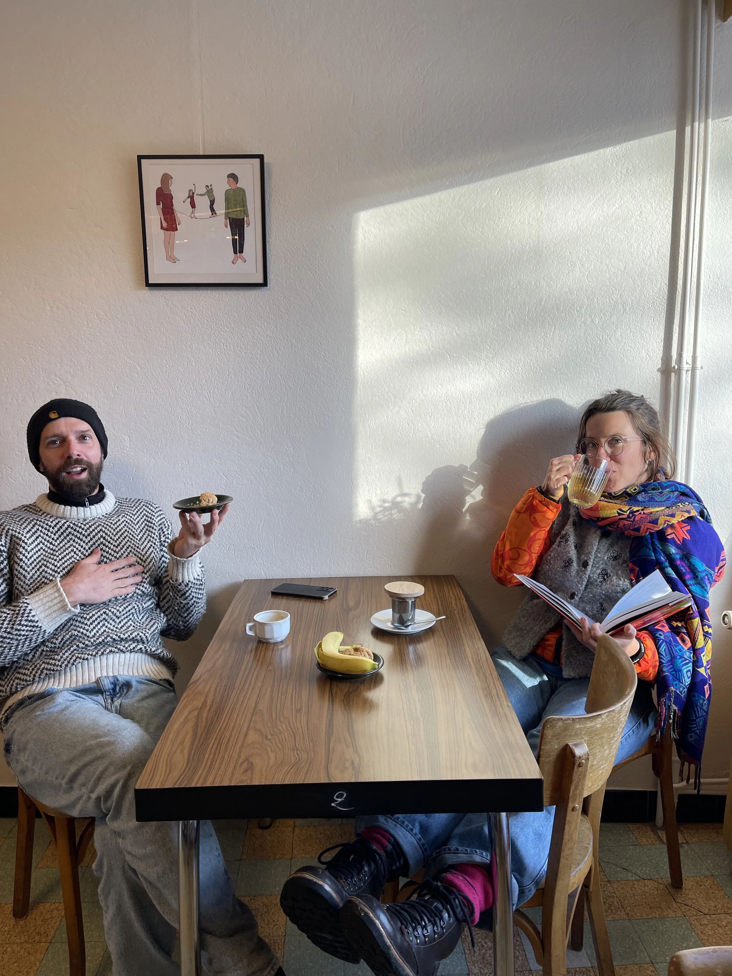 Deux personnes assises à une table en bois dans une pièce, l'un tient une tasse et l'autre lit un livre tout en buvant. La table a une banane, une tasse, une assiette avec un biscuit, une petite jarre en céramique, et un téléphone. Un tableau avec de