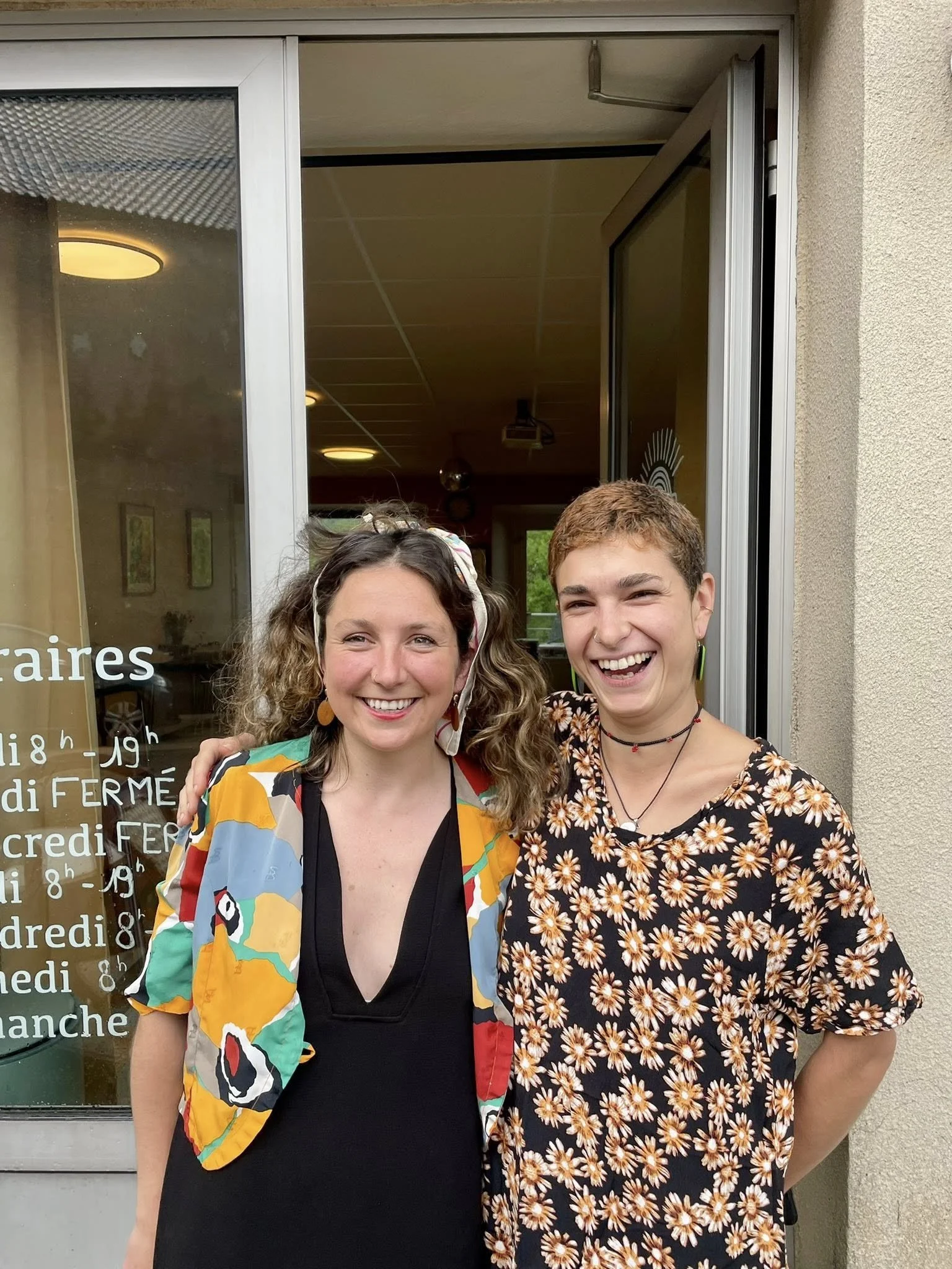Deux jeunes femmes souriantes posant devant une porte d'entrée d'une boulangerie. l'une porte une robe noire avec une veste colorée, l'autre porte une robe à fleurs